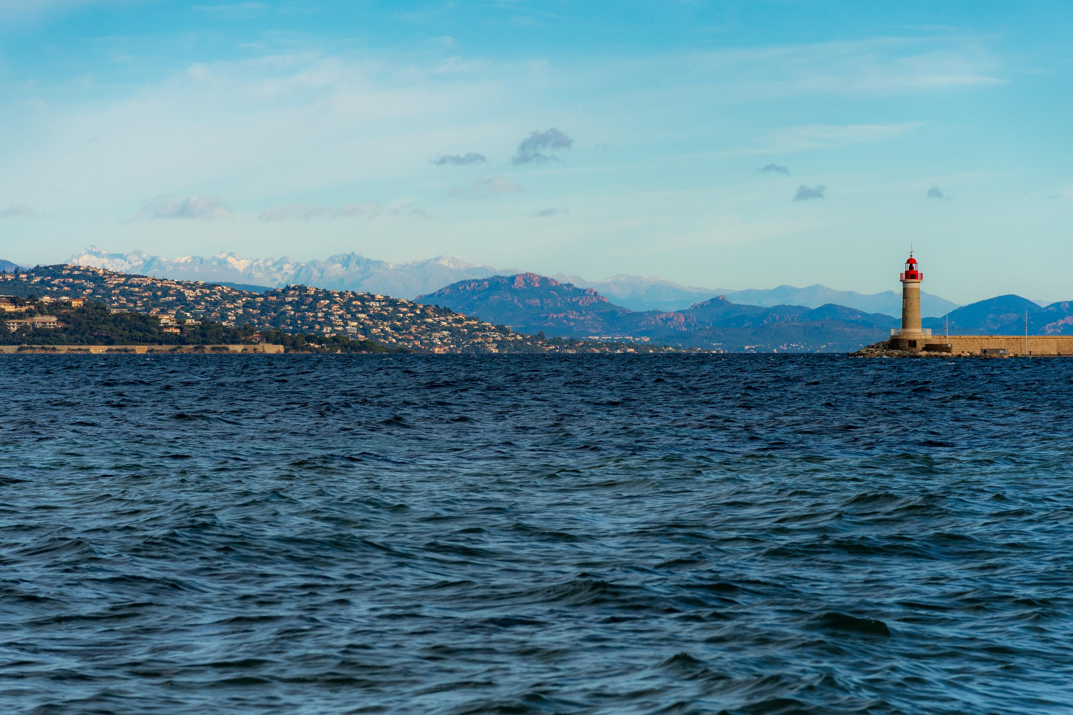 Das Foto zeigt eine ruhige Meereslandschaft mit einem Leuchtturm im Vordergrund. Das tiefe Blau des Wassers bildet einen starken Kontrast zum hellen Himmel, der von einigen Wolken durchzogen ist. Im Hintergrund erstreckt sich eine Küstenlinie mit dicht bebauten Hügeln und dahinter aufragenden Bergen, deren Gipfel leicht mit Schnee bedeckt sind. Der Leuchtturm steht auf einem steinernen Damm und scheint als Orientierungspunkt für Schiffe zu dienen. Die Szene strahlt eine friedliche und leicht melancholische Stimmung aus, die durch das weite Meer und die ferne Bergkulisse verstärkt wird. Es ist ein Bild, das an die Weite und die Schönheit der Natur erinnert und gleichzeitig die menschliche Präsenz durch die Bebauung und den Leuchtturm andeutet.