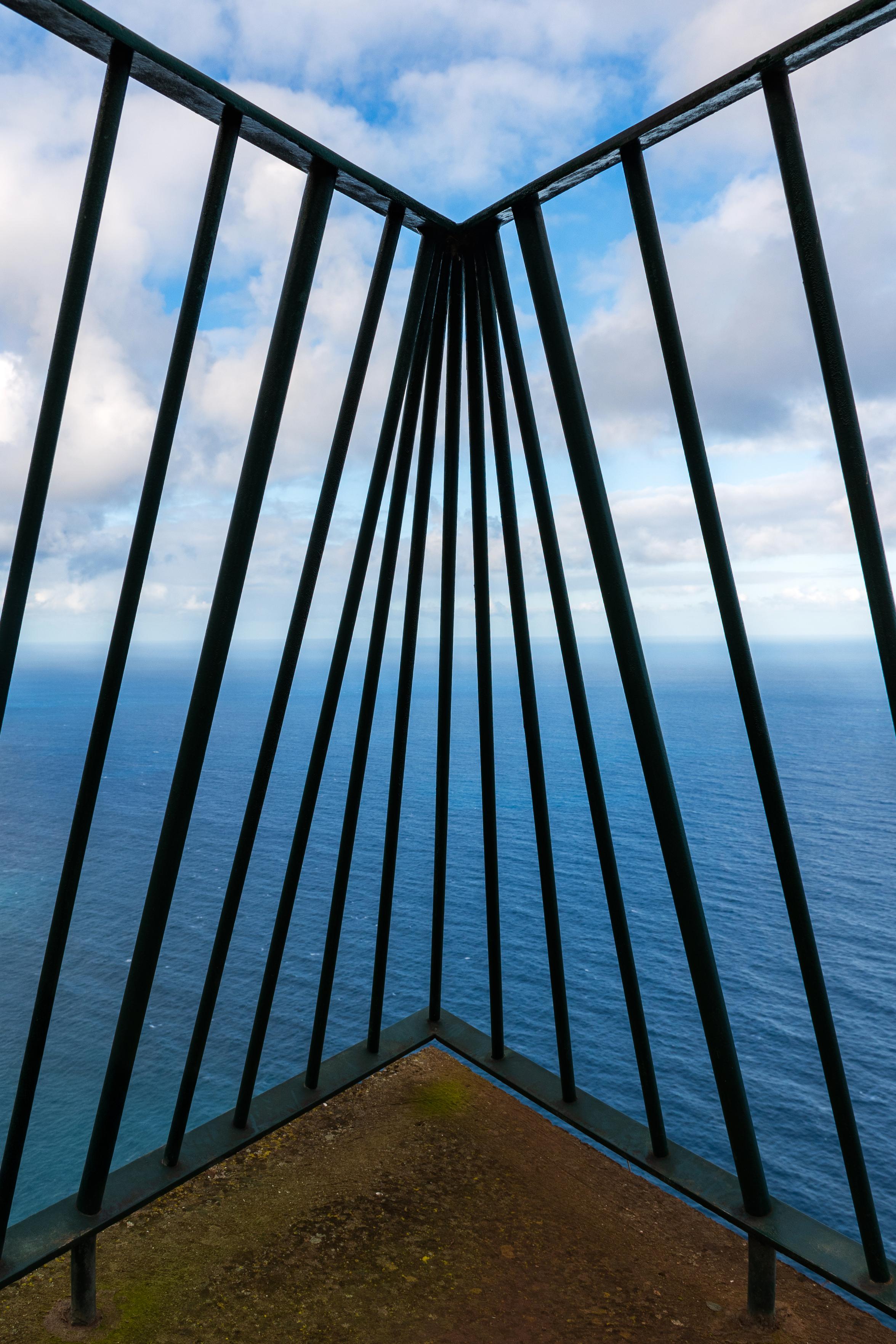 Das Foto zeigt eine Perspektive von einem Aussichtspunkt, der von einer Metallkonstruktion eingerahmt wird. Die dunklen, vertikalen Streben bilden einen Rahmen, der den Blick auf das weite, blaue Meer und den Himmel freigibt. Die Wolken am Himmel sind leicht und flauschig, und das Meer ist ruhig, aber mit einer leichten Wellenbewegung. Der Boden unter den Füßen ist rau und mit Moos bewachsen.