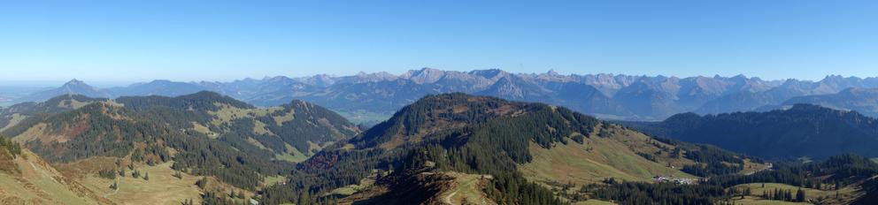 Panoramabild von einem Gipfel des Allgäus auf die ganze südliche Gebirgskette Westtirol. Himmel über den nicht schneebedeckten Gipfeln ist strahlendblau in Mittagssonne. Im Vordergrund Berge mit Nadelwäldern und grünen Wiesen, die sich abwechseln.