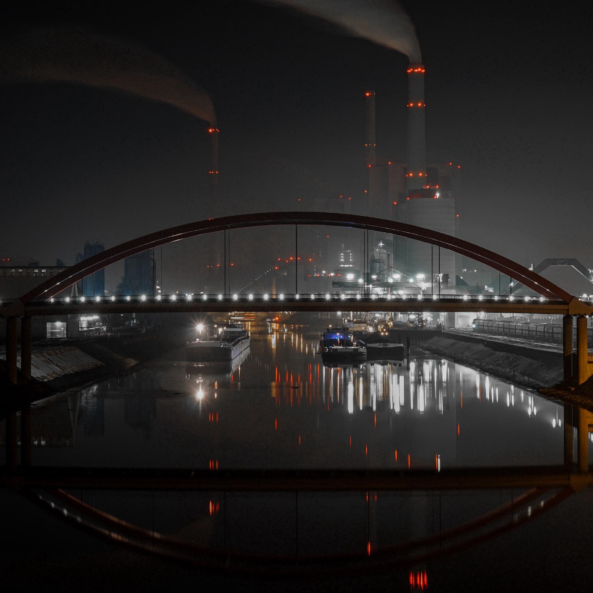 Im Vordergrund die Bogenbrücke der Duisburger Straße mit ihrer perlenschnurartigen Beleuchtung über das dunkle Hafenbecken hinweg, dahinter die hohen Kesselbauten und Schlote des Mannheimer Großkraftwerks, mit seinen vielen Lichtern, alles überdacht von einem dunklen Nachthimmel.