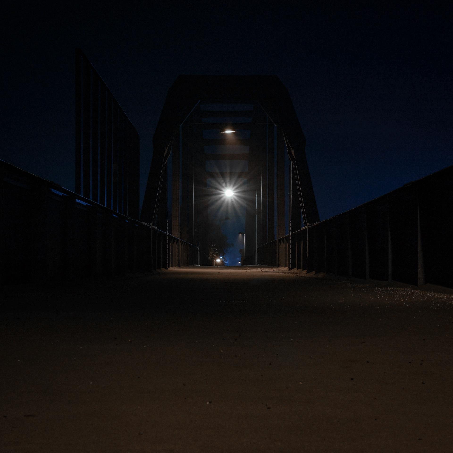 Eine bogenförmige Fußgängerbrücke, der sogenannte Graßmannsteg, über das Hafenbecken gespannt, mit einer einsamen, weißen Leuchte gebau in der Mitte, unter einem dunklen Nachthimmel.
