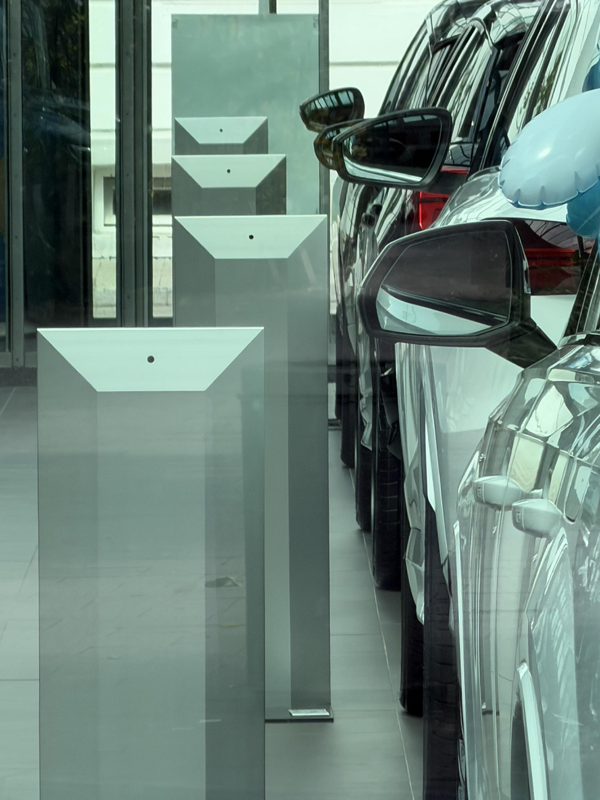 Eine Reihe von Autos steht dicht hintereinander in einem Ausstellungsraum. Ihre Seitenspiegel wiederholen sich bis in den Hintergrund, während links parallel dazu helle geometrische Ausstellungselemente in die Tiefe führen.