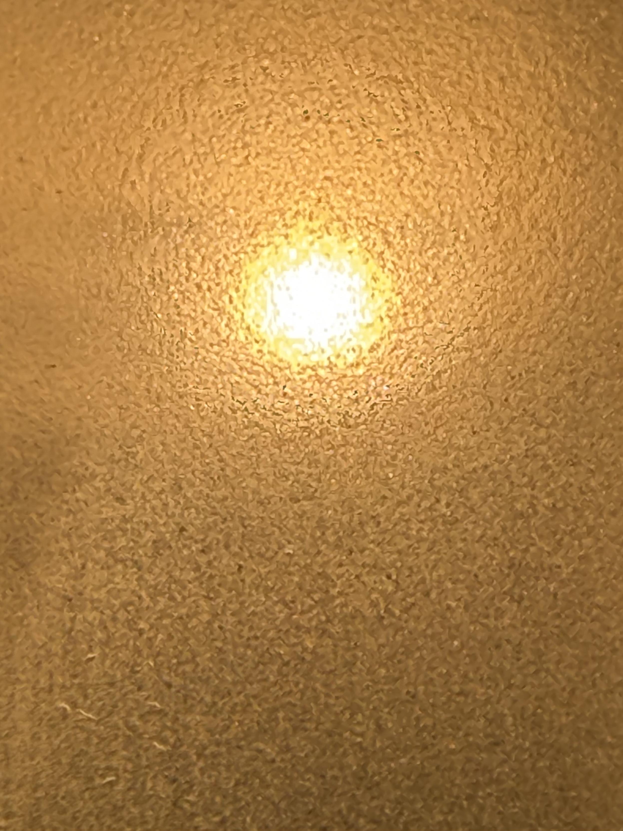 Nahaufnahme einer orange-gelben, strukturierten Oberfläche mit einem hell leuchtenden Zentrum. Das Licht scheint sich durch die körnige, reliefartige Materie nach außen zu drücken und erzeugt einen dichten, glühenden Eindruck.
