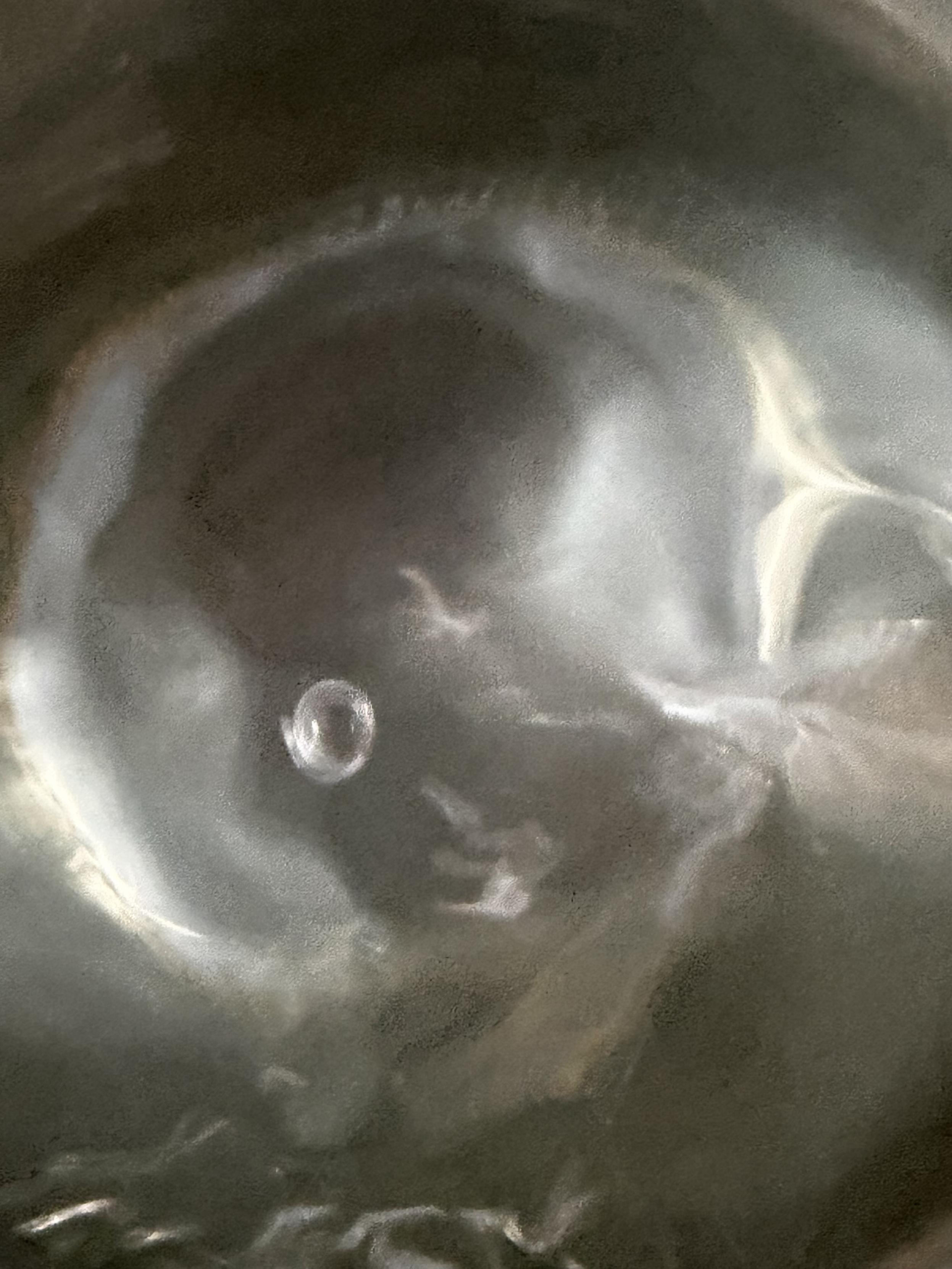 Nahaufnahme von Wasser, das sich um eine rundliche, blasenartige Form sammelt. Licht und Schatten bewegen sich über die dunkle Oberfläche, während das Wasser für einen kurzen Moment eine durchscheinende Form hält.
