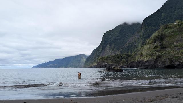 Schwarzer Sandstrand, an den kleinen Wellen schlagen. Hüfthoch im Wasser stehend ein engumschlungendes Paar, im Hintergrund grüne Klippen. Himmel grau und bedeckt.