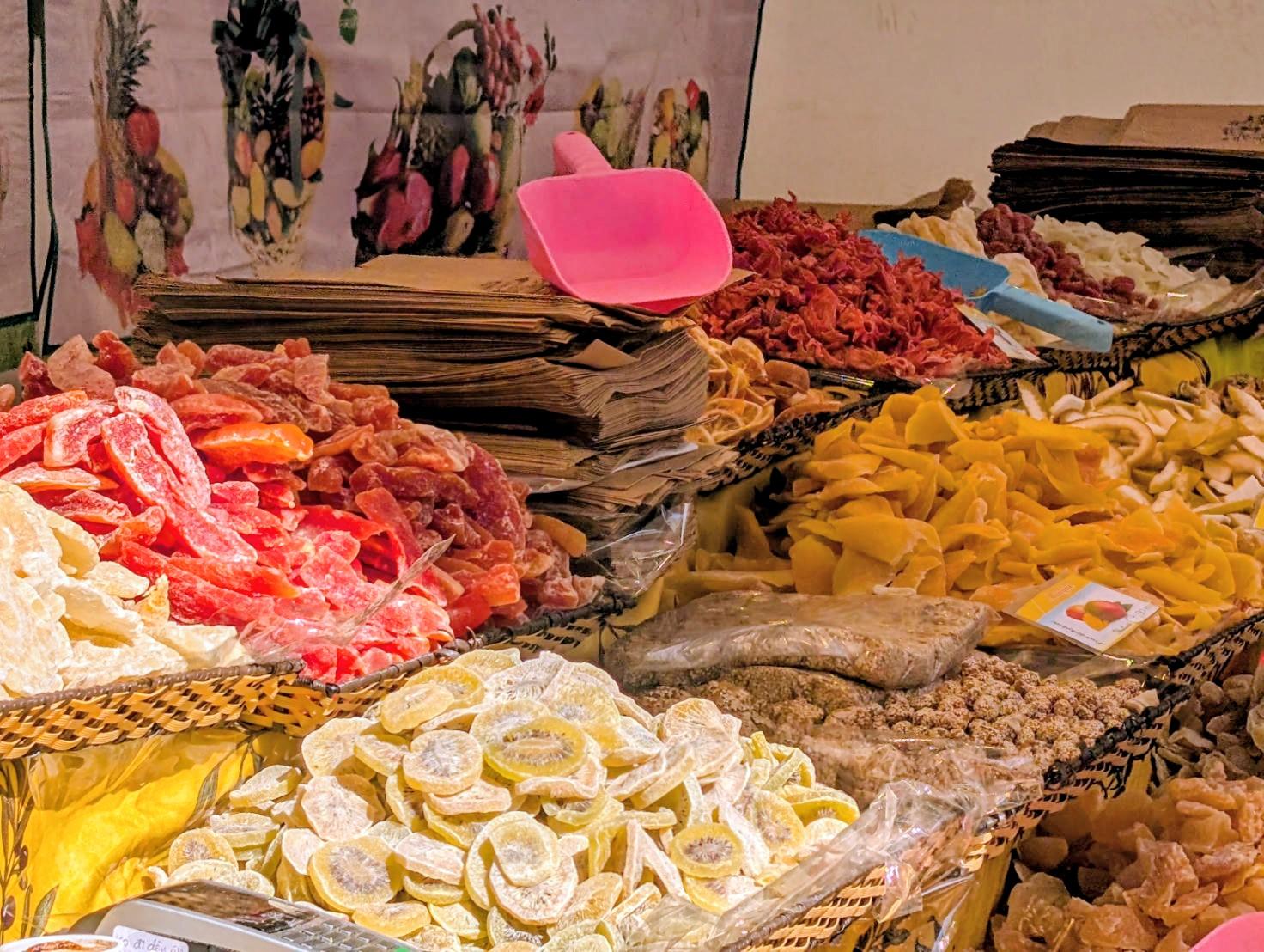 Ein Stand mit Bergen von getrockneten Früchten. Im Vordergrund sind getrocknete Kiwi, Ingwer und Papaya zu sehen, dazwischen braune Papertüten und eine große rote Plastikschaufel. Im Hintergrund getrocknete Mango und andere Früchte