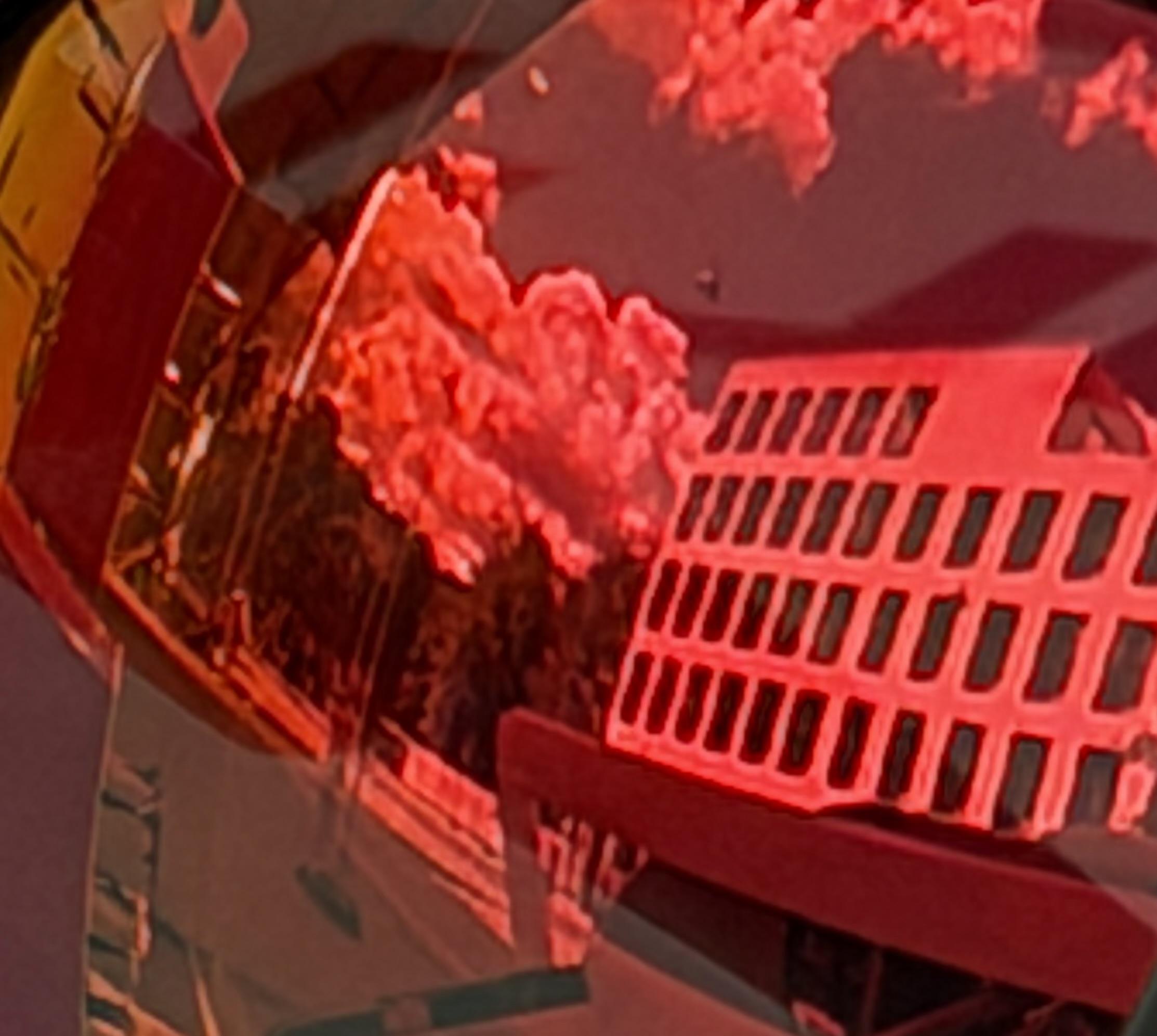 Eine leuchtend rote urbane Spiegelung füllt das Bild. Rechts erscheint ein mehrstöckiges Gebäude mit Reihen dunkler Fenster, während sich Bäume, Wolken, Straßenstrukturen und Fragmente der umgebenden Architektur auf einer gewölbten spiegelnden Fläche überlagern