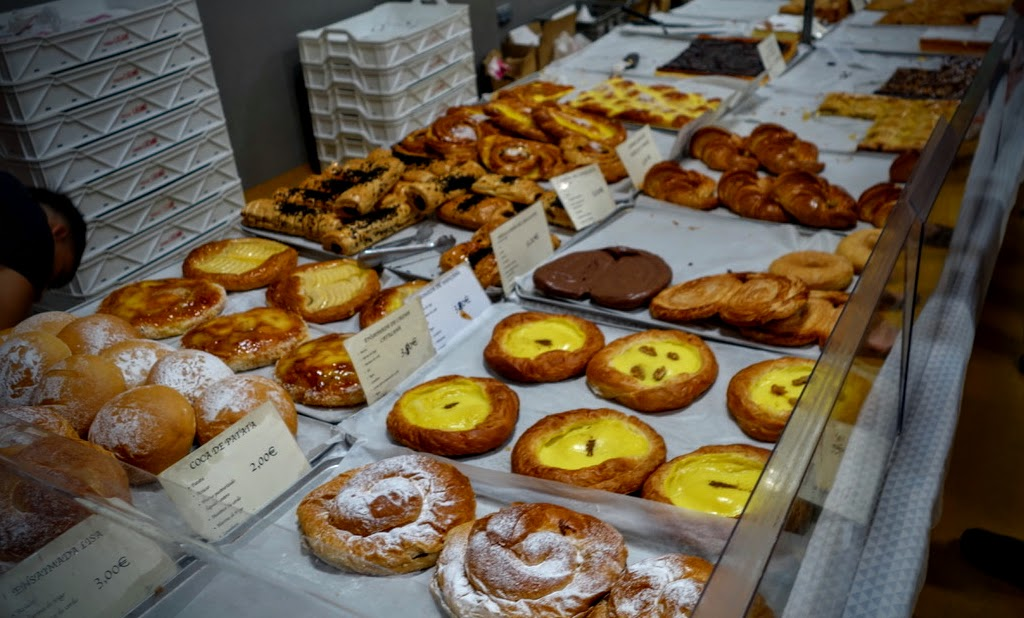 Verkaufstheke mit zahlreichen süßen und herzhaften Backwaren auf Blechen. Zu sehen sind unter anderem portugiesische Puddingtörtchen mit gelber Creme, Croissants, Blätterteiggebäck, Schokogebäck und große, mit Puderzucker bestäubte Schnecken. Daneben stehen kleine Preisschilder, im Hintergrund stapeln sich weiße Transportkisten.