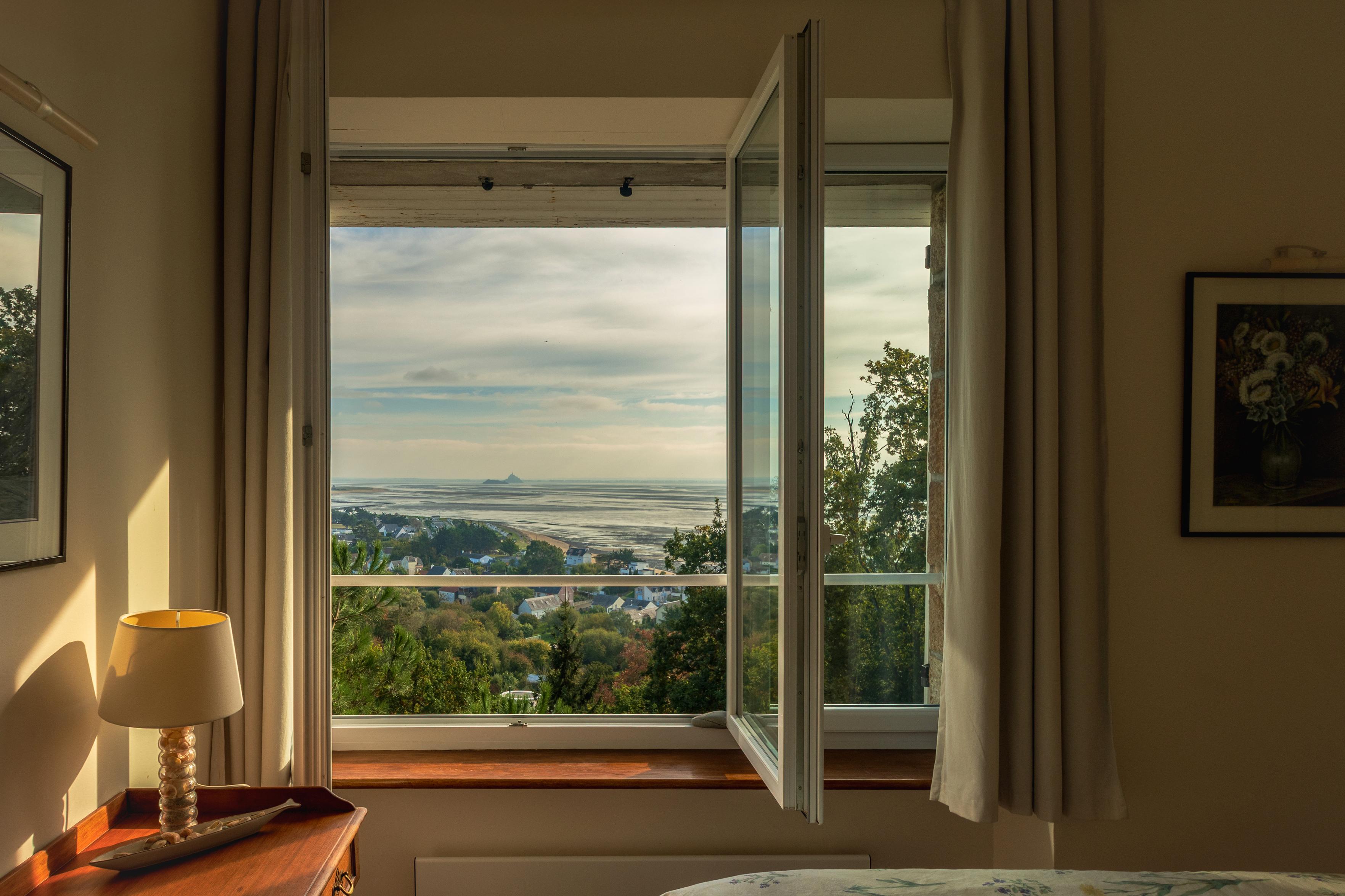 Das Bild zeigt einen Blick aus einem offenen Fenster auf eine Küstenlandschaft. Im Vordergrund steht ein Nachttisch mit einer Lampe, die von der tiefstehenden Sonne angestrahlt wird. Durch das Fenster sieht man ein kleines Dorf, das sich an einen Hang schmiegt, umgeben von üppigem Grün. Weiter hinten erstreckt sich eine Bucht bei Ebbe bis zum Horizont, wo man schemenhaft den berühmten Mont Saint Michel erkennen kann. Der Himmel ist von Wolken durchzogen, die das Licht der Sonne brechen und eine friedliche, leicht melancholische Atmosphäre schaffen. Die Szene vermittelt ein Gefühl von Ruhe und Geborgenheit, während gleichzeitig die Weite und Schönheit der Natur zum Träumen einladen. Es ist ein Moment des Innehaltens, der die Verbindung zwischen dem Innenraum und der Außenwelt aufzeigt.