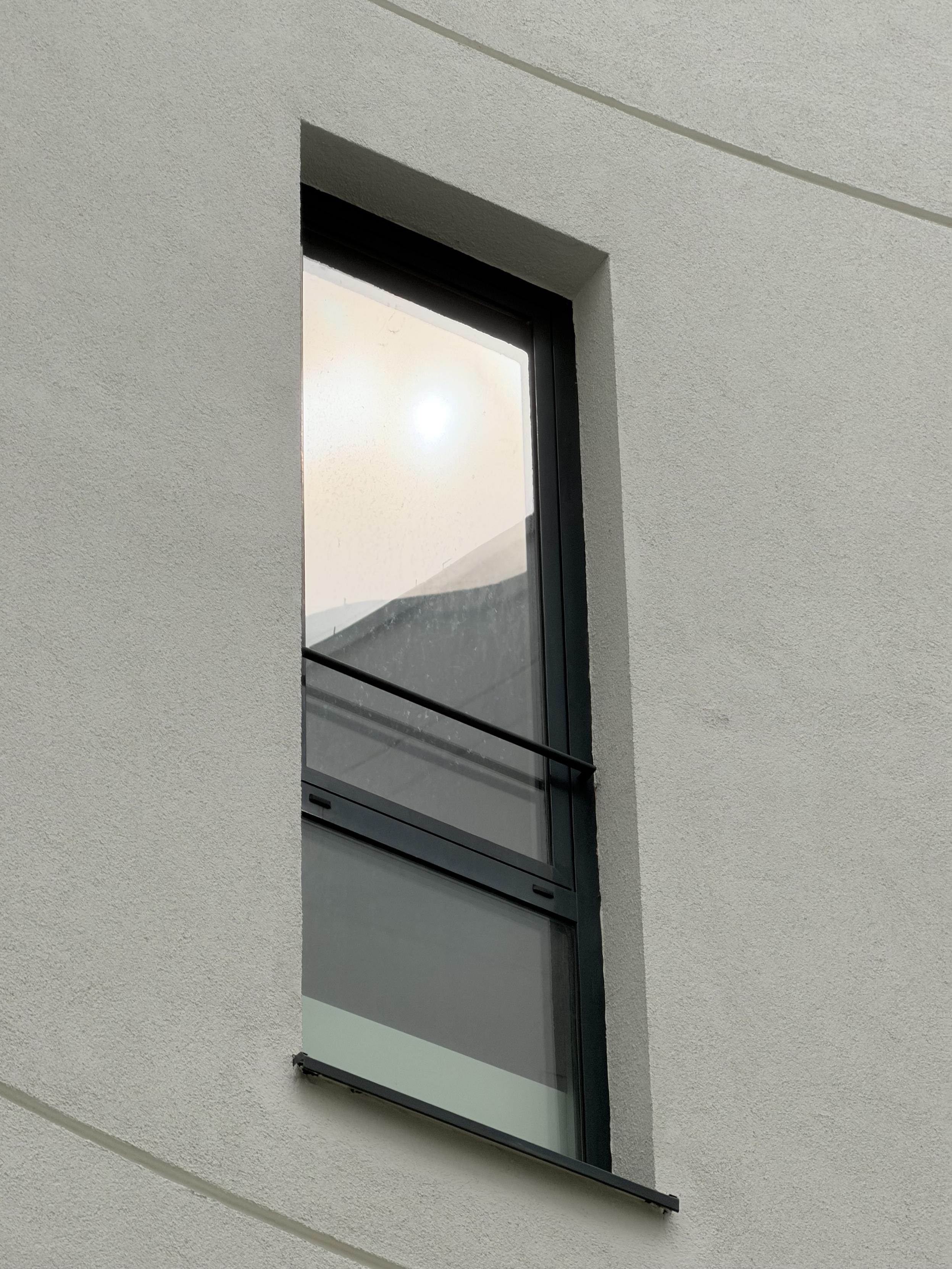 Ein rechteckiges Fenster sitzt hoch in einer sich rundenden, hellen Wand und spiegelt Konturen unter einem hellen Himmeli. Die umgebende Wand verläuft sanft geschwungen und lässt den größten Teil des Bildes still und leer.