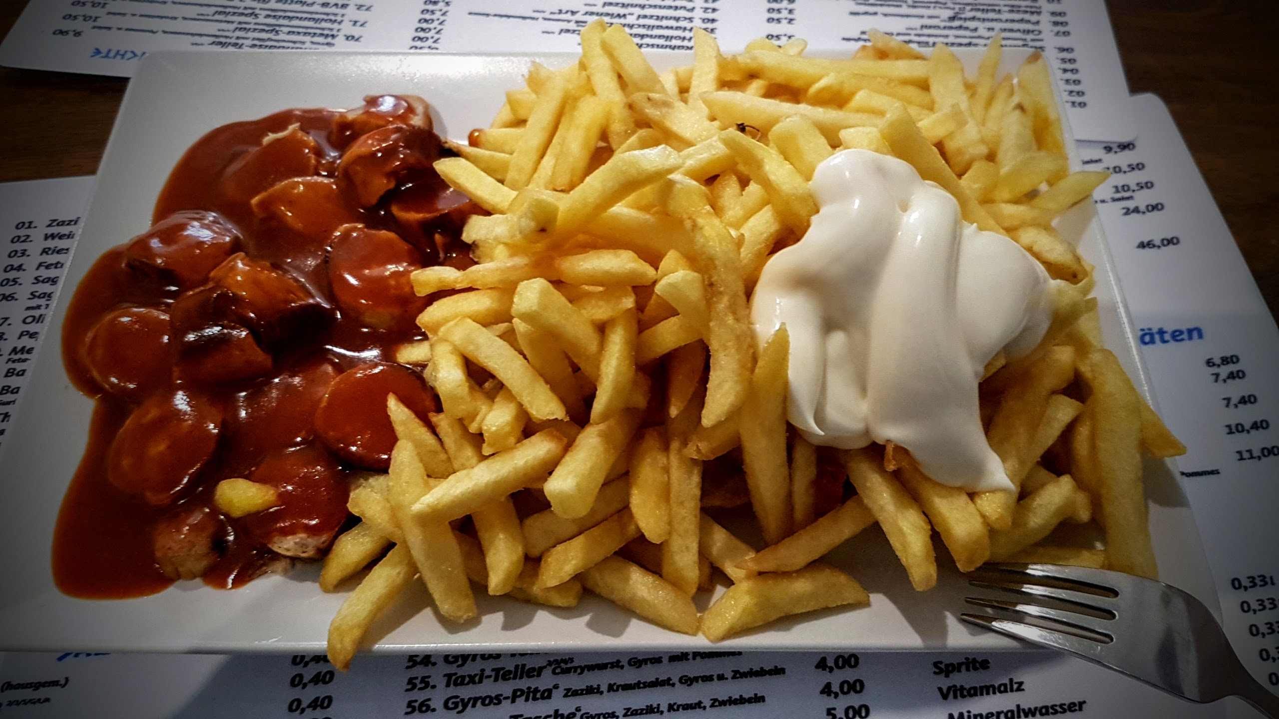 Ein Teller mit einer großen Portion goldbrauner Pommes frites. Daneben liegen mehrere Stücke Wurst in einer dunkelroten Soße. Auf den Pommes befindet sich ein großer Klecks weißer Mayonnaise. Rechts unten liegt eine Gabel. Der Teller steht auf einer Speisekarte.