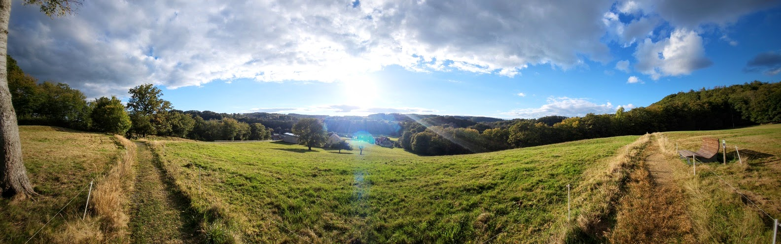 Panoramaartige, durch die Weitwinkelaufnahme leicht verzerrte Landschaft: Ein schmaler Grasweg verläuft bogenförmig von links über die Wiese nach rechts und ist an beiden Enden des Bildes zu sehen. Die weitläufige, hügelige Wiese ist von einzelnen Bäumen und bewaldeten Hügeln umgeben. Im Hintergrund sind einige Häuser zu erkennen. Tief stehende Sonne scheint durch eine Wolkenlücke und erzeugt einen hellen Lichtschein und eine leichte Lens-Flare-Spur. Darüber ein weiter Himmel mit dichten Wolken und blauen Partien.