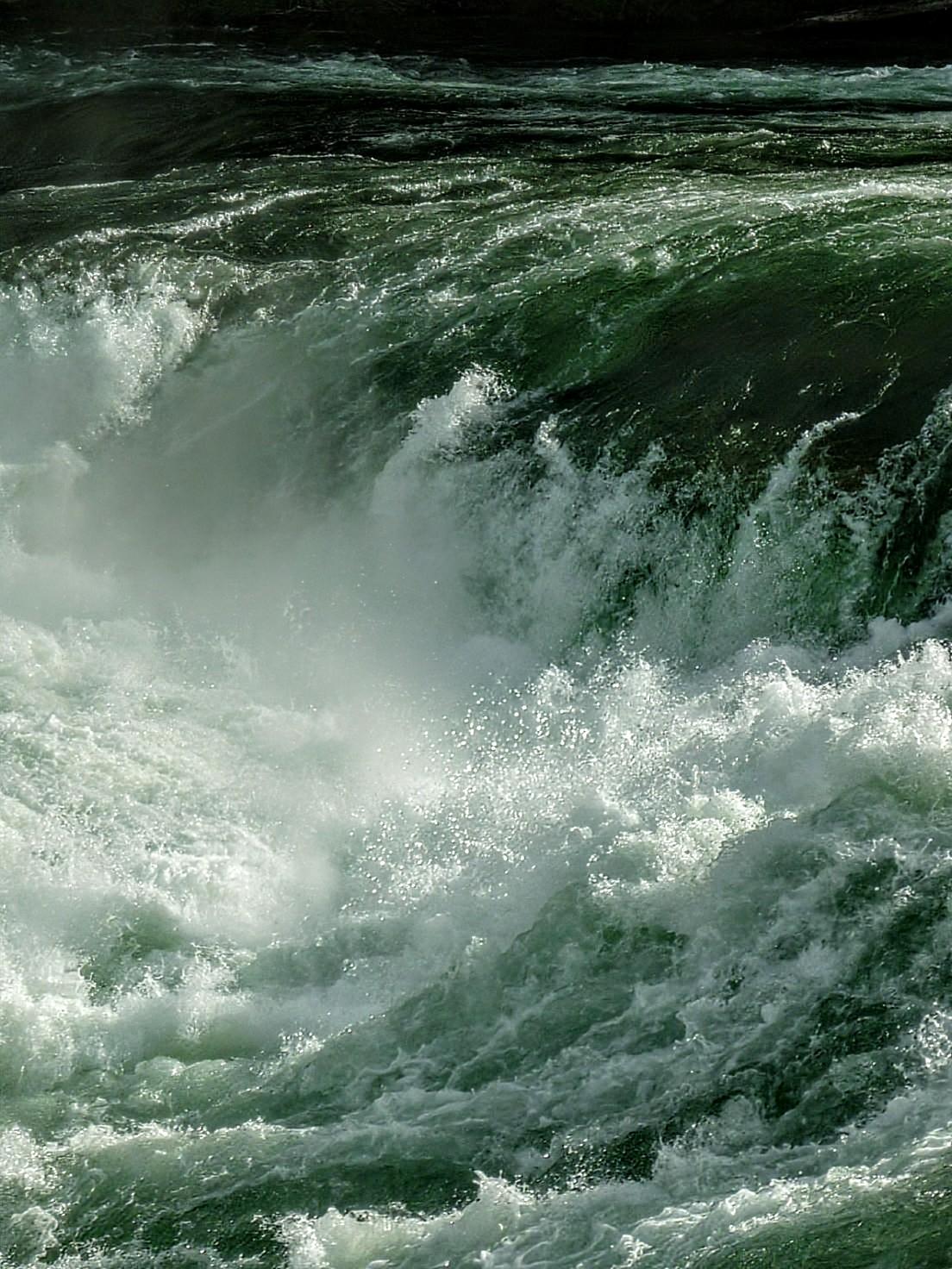 Nahaufnahme einer Stromschnelle in einem Fluss. Oben rechts strömt das Wasser geradeaus, verwirbelt sich dann in der Mitte des Bildes. Die spritzende Gischt und einzelne Tropfen sind zu sehen.