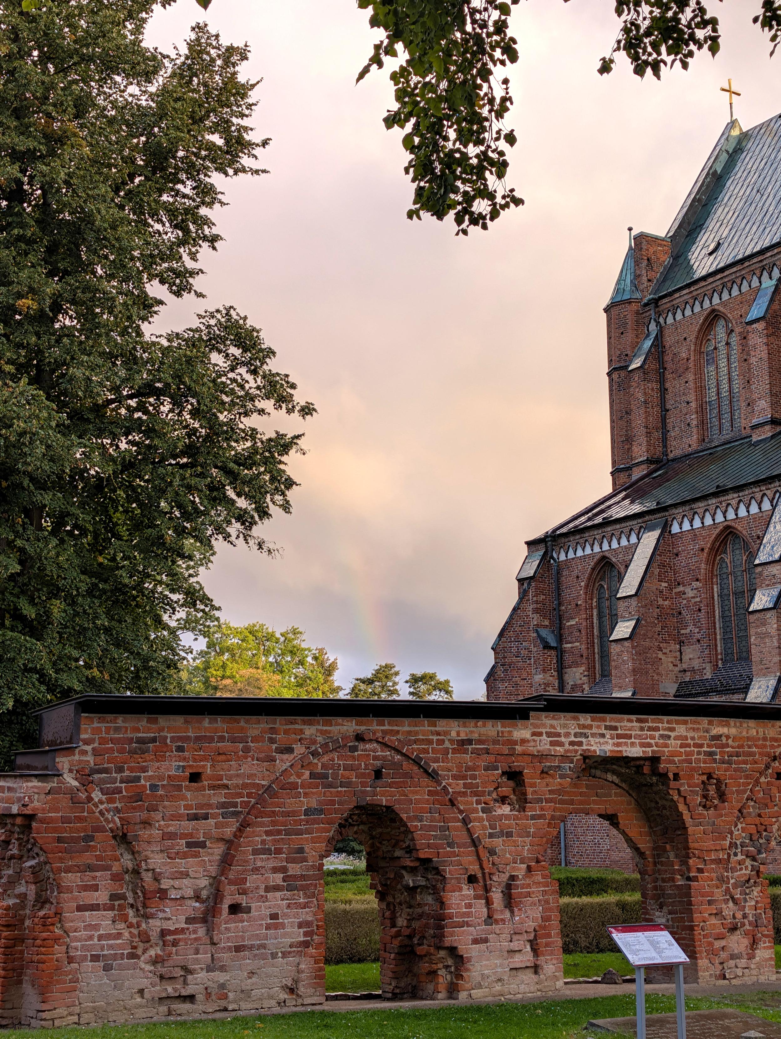 Der Betrachter steht vor einer mittelalterlichen Backsteinmauer mit Rundbogenöffnungen. Rechts dahinter sind das westliche Langhaus und das südliche Seitenschiff einer großen Backsteinkirche mit nassen Kupferdächern zu erkennen. Links sieht man einen großen Laubbaum. Einige Zweige hängen auch von oben ins Bild. Der in der Bildmitte sichtbare Himmel ist bewölkt. Ein Regenbogen ist auszumachen.