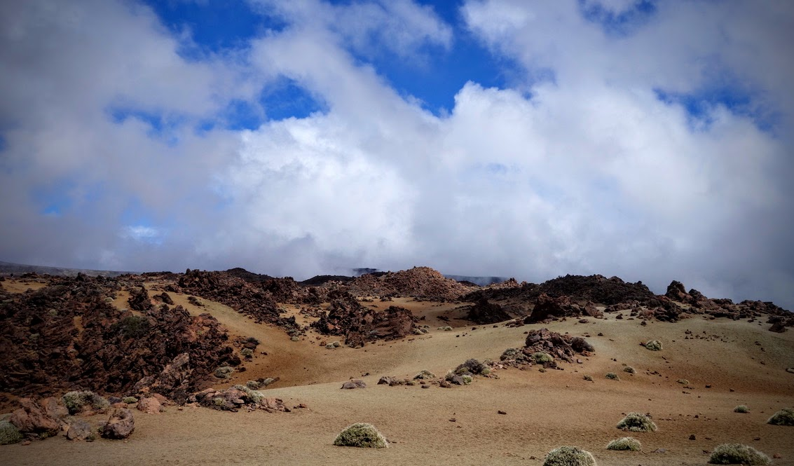 Karge, vulkanische Landschaft unter einem dramatischen Himmel. Eine weite, sandige Ebene ist von dunklen rotbraunen Lavasteinen, Geröll und niedrigen, rundlichen Pflanzen übersät. Im Hintergrund erheben sich flache Geröllhügel. Darüber ziehen dichte weiße und graue Wolken auf, zwischen denen stellenweise kräftig blauer Himmel sichtbar ist.