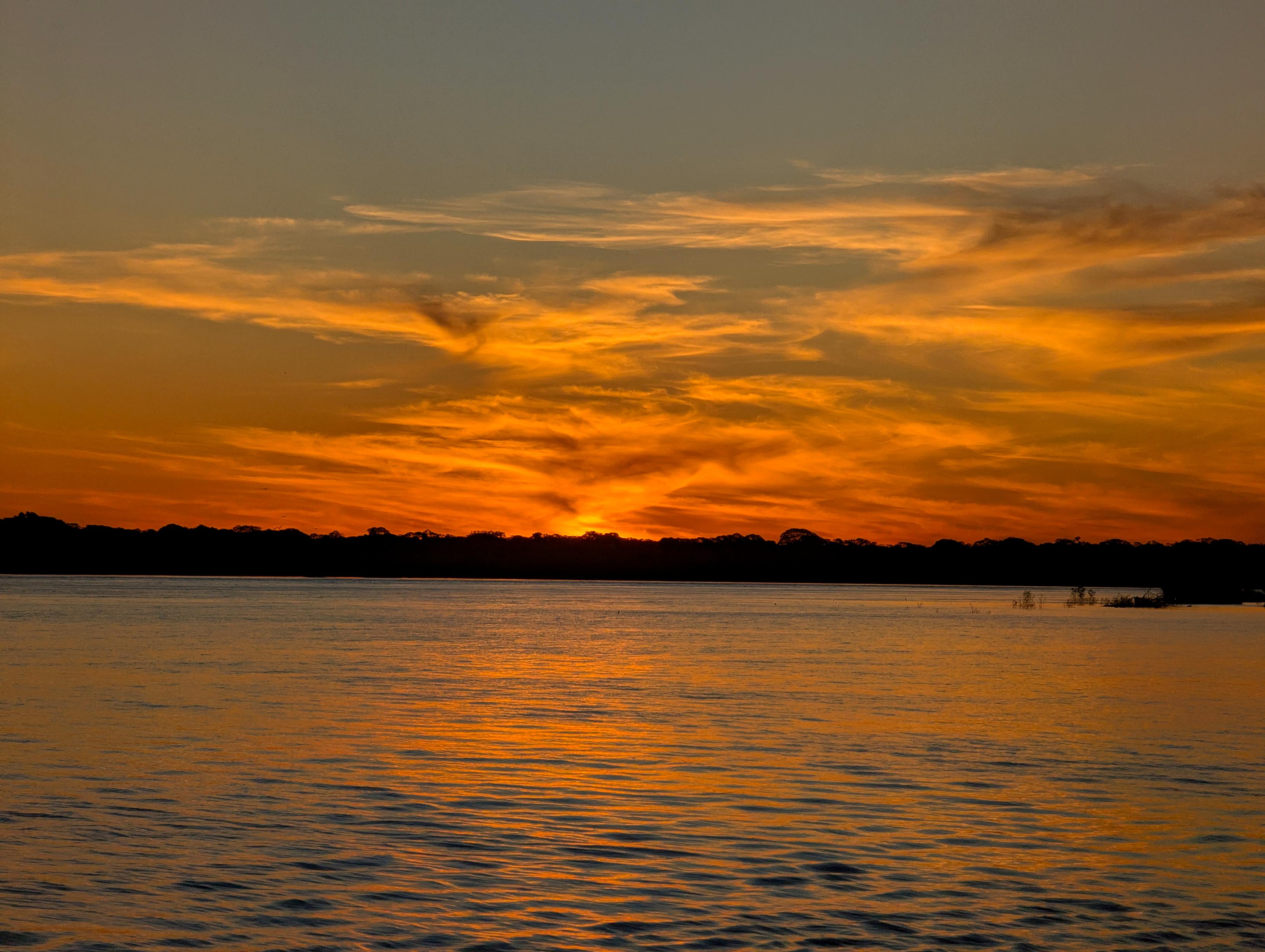 Der Betrachter blickt über die leicht gekräuselte große Wasserfläche eines Flusses in den Sonnenuntergang. Lockere Wolken um die Sonne herum sind orange und goldgelb gefärbt. Ein dichter Wald unterhalb ist als dunkle Silhouette am jenseitigen Ufer zu erkennen. Das Wasser davor erscheint matt grau mit orangefarbenem Glanz.