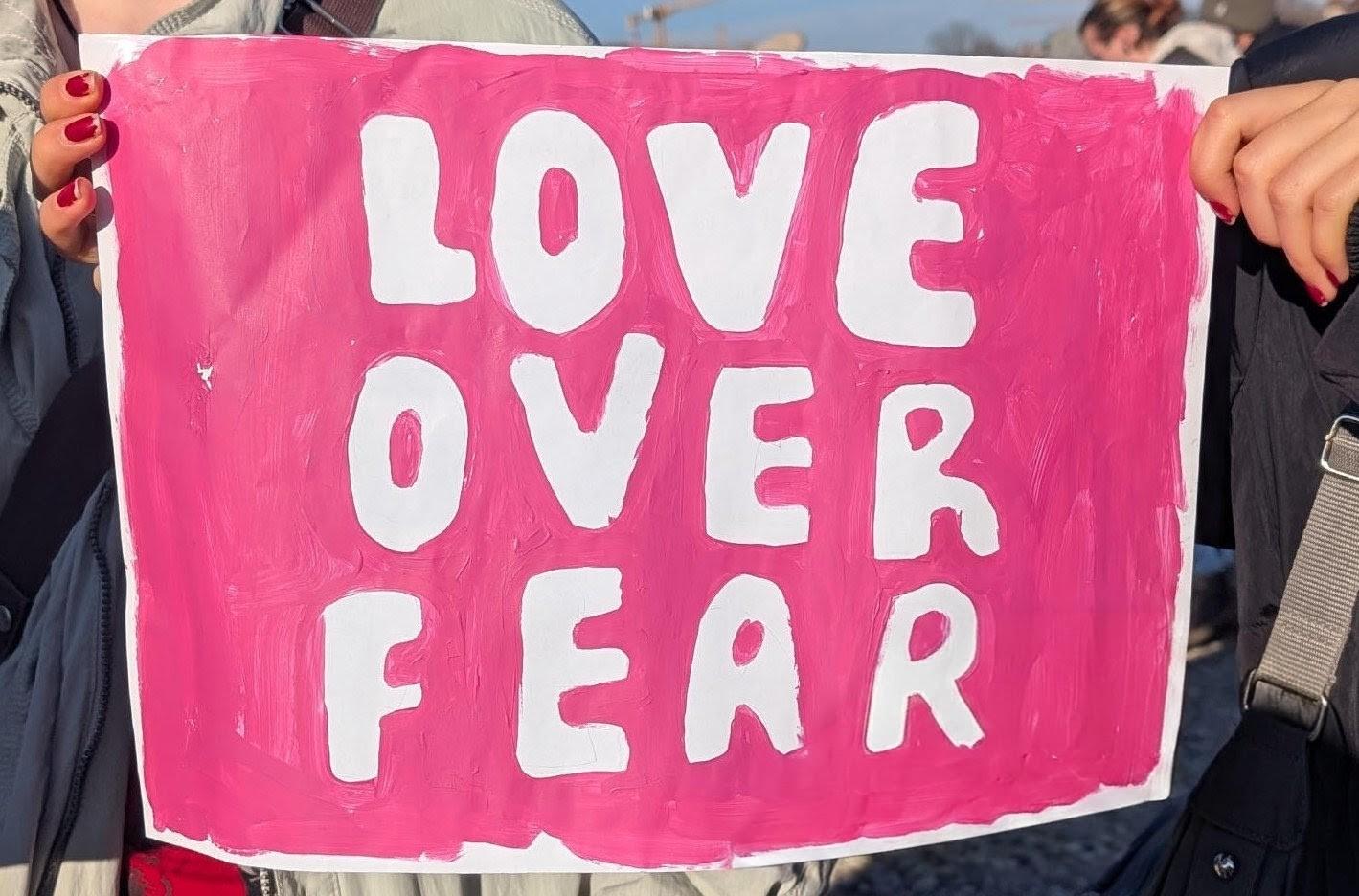 Ein Pappschild auf einer Demo. Darauf steht "LOVE OVER FEAR" 
Das Schild ist pink, die Schrift weiß.Es wird links und rechts von zwei Personen gehalten. Nur deren Hände sind zu sehen, die Fingernägel rot lackiert.