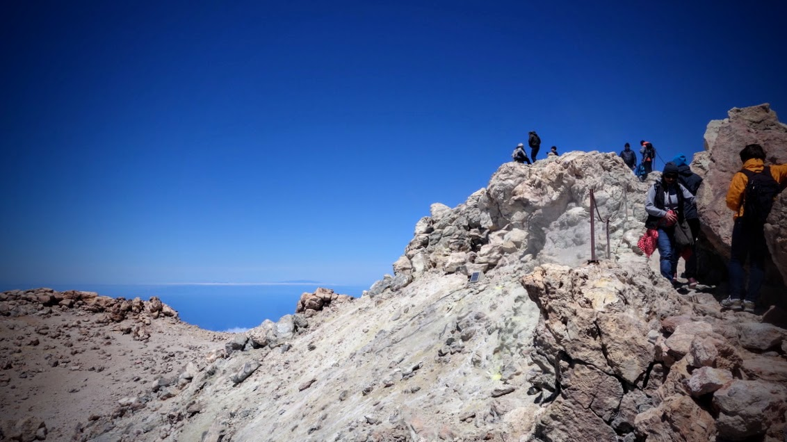 Menschen stehen und klettern auf einem felsigen Berggipfel. Im Hintergrund erstrecken sich unter einem tiefblauen, wolkenlosen Himmel Meer und Horizont. Die felsige Landschaft ist hell und karg; rechts im Bild sind mehrere Wandernde mit Rucksäcken zu sehen.