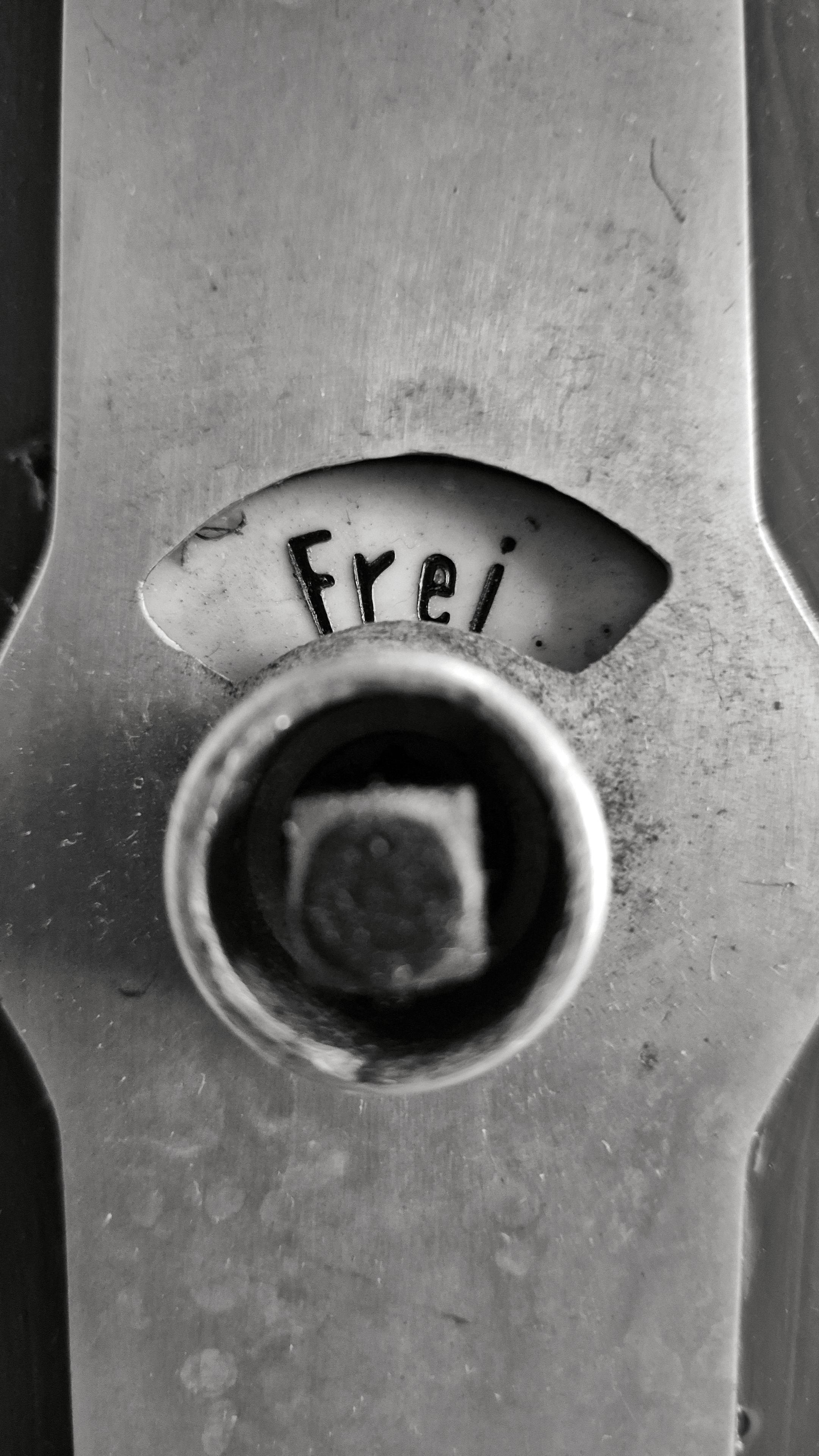 Das Foto ist rechteckig und vertikal ausgerichtet. Es ist schwarz-weiß. Zu sehen ist eine Nahaufnahme eines Teils eines metallenen Türbeschlags einer Toilettentür aus den 1960er Jahren. In einem gebogenen Ausschnitt des Türbeschlags oben im Bild steht in dunklen Buchstaben auf hellem Grund die Aufschrift „Frei“. Darunter befindet sich ein runder Metallkreis mit einer quadratischen Nuss in der Mitte.