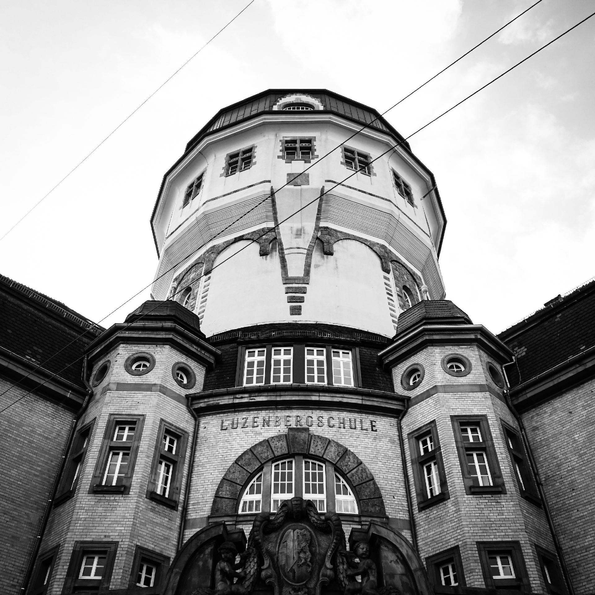 Ein wuchtiger Wasserturm erhebt sich rund in den Himmel, drumherum die Schulgebäude. Das Fot ist schwarzweiß.