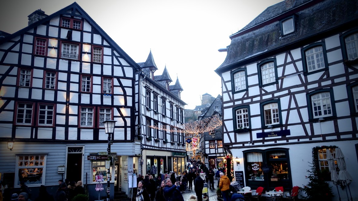 Historische, dicht bebaute Altstadtgasse mit mehreren schwarz-weißen Fachwerkhäusern. Links ein großes, mehrstöckiges Fachwerkhaus mit roten Fensterrahmen, rechts ein weiteres großes Fachwerkhaus. In der Mitte führt die schmale, belebte Straße bergauf; zahlreiche Menschen sind unterwegs. Im Hintergrund sind weitere Fachwerkhäuser und ein Turm auf einem Hügel zu sehen. Die Aufnahme entstand in der bläulichen Dämmerung.