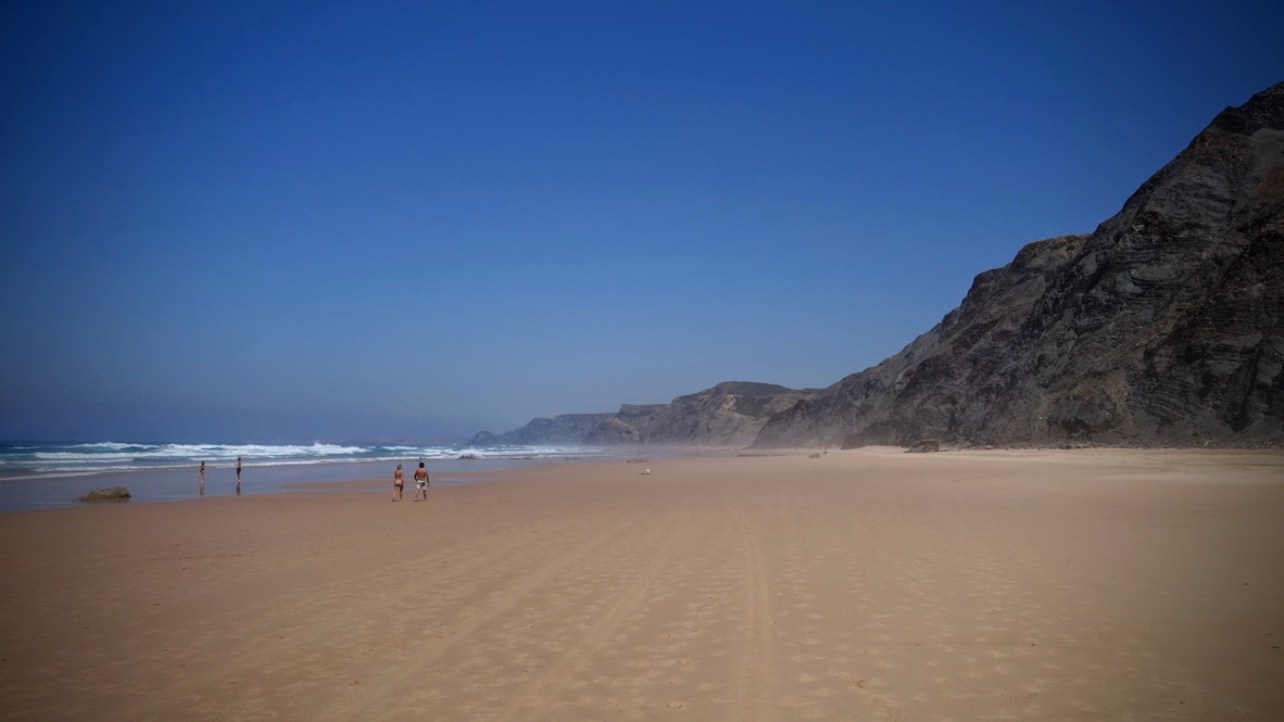 Weitläufiger, fast menschenleerer Sandstrand unter tiefblauem Himmel. Links am Bildrand liegt das Meer mit sanften Wellen. Im flachen Wasser und am Strand stehen vier kleine Personen. Rechts erhebt sich eine steile, dunkle Felswand, die sich weit in die Ferne entlang der Küste zieht. Der feine Sand ist von leichten Spuren durchzogen. Im Hintergrund liegt die Küste in einem bläulichen Dunst.