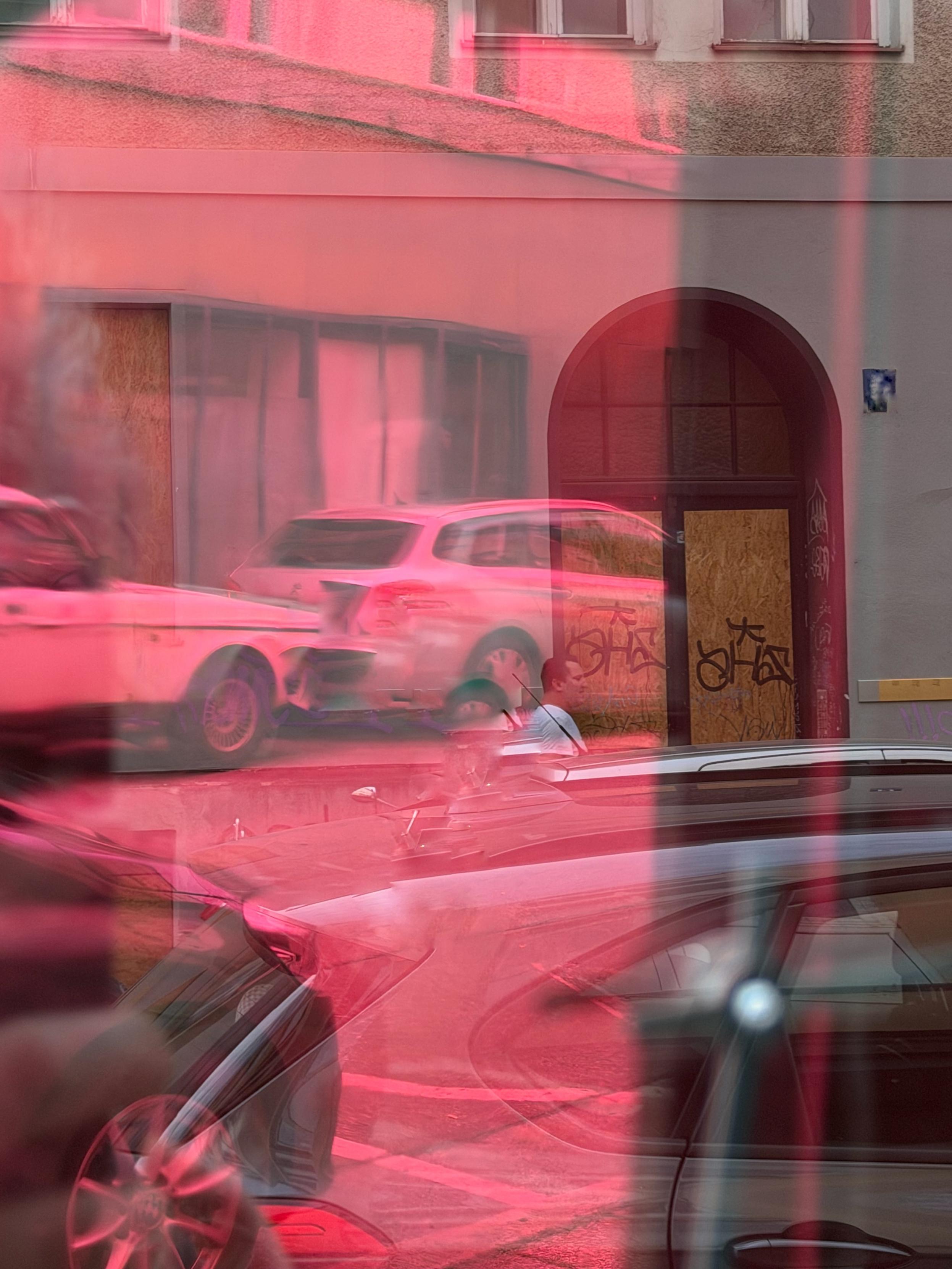 Straßenszene, stark in Pink- und Rottönen überlagert, durch eine spiegelnde oder getönte Scheibe fotografiert. Im Vordergrund verläuft die glänzende Motorhaube und Dachlinie eines dunklen Autos, an der sich weitere Fahrzeuge und die Straße spiegeln. Dahinter eine Häuserfassade mit einem gewölbten Torbogen, dessen Holztür großflächig mit Graffiti-Schriftzügen bedeckt ist. Rechts und links parken mehrere Autos, eine Person in weißem T-Shirt steht zwischen den Fahrzeugen. Ein durchgehender vertikaler dunkler Streifen zieht sich über die gesamte Bildhöhe.