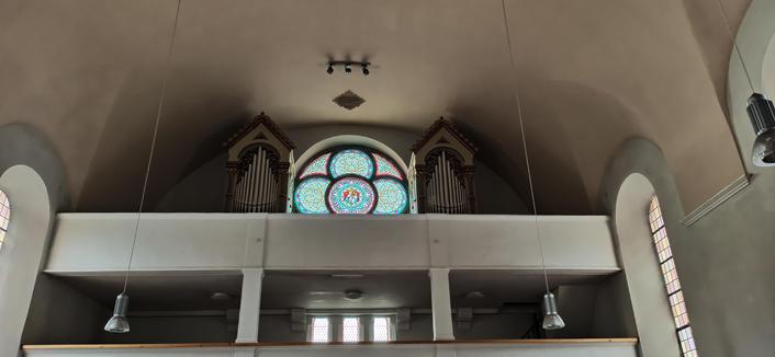 AUf ener doppelstöckigen Empore einer Lirche steht  eine Orgel, verteilt auf zwei Seiten links und rechts eines runden Fensters. Das Fenster ist mit vier sich überschneidenden kreisrunden Fensterrahmen unterteilt und mit bunten Glasmotiven versehen. Das Licht, das duc hdie Scheibe fällt, läßt die Farben erstrahlen.