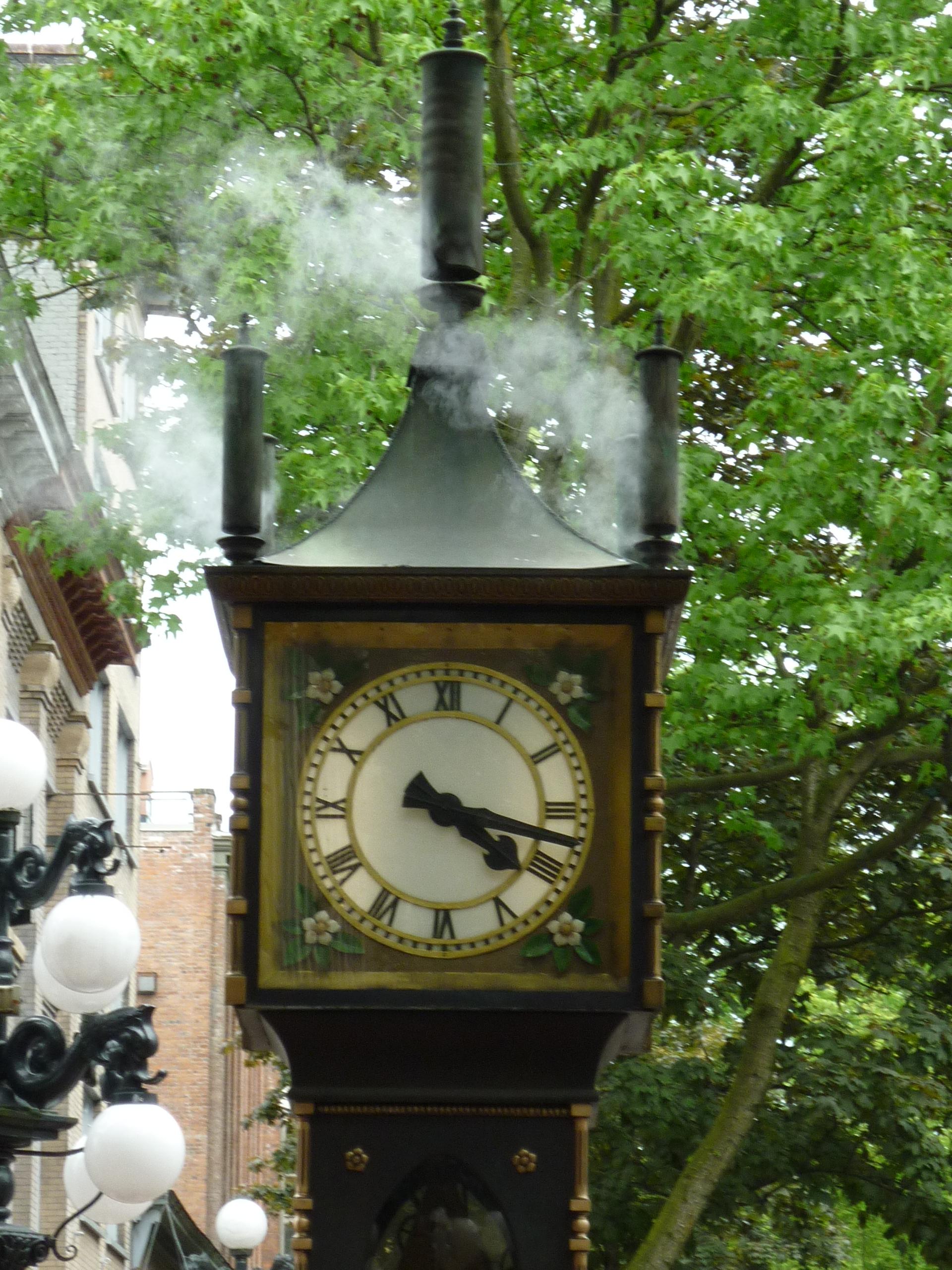 Eine rauchende Uhr in der Altstadt. Ein historisches Ziffernblatt mit römischen Zahlen, ein spitzes Dach und oben auf dem Dach drei Pfeifen, aus denen Dampf strömt. Im Hintergrund ein grün belaubter Baum, links ein Haus.