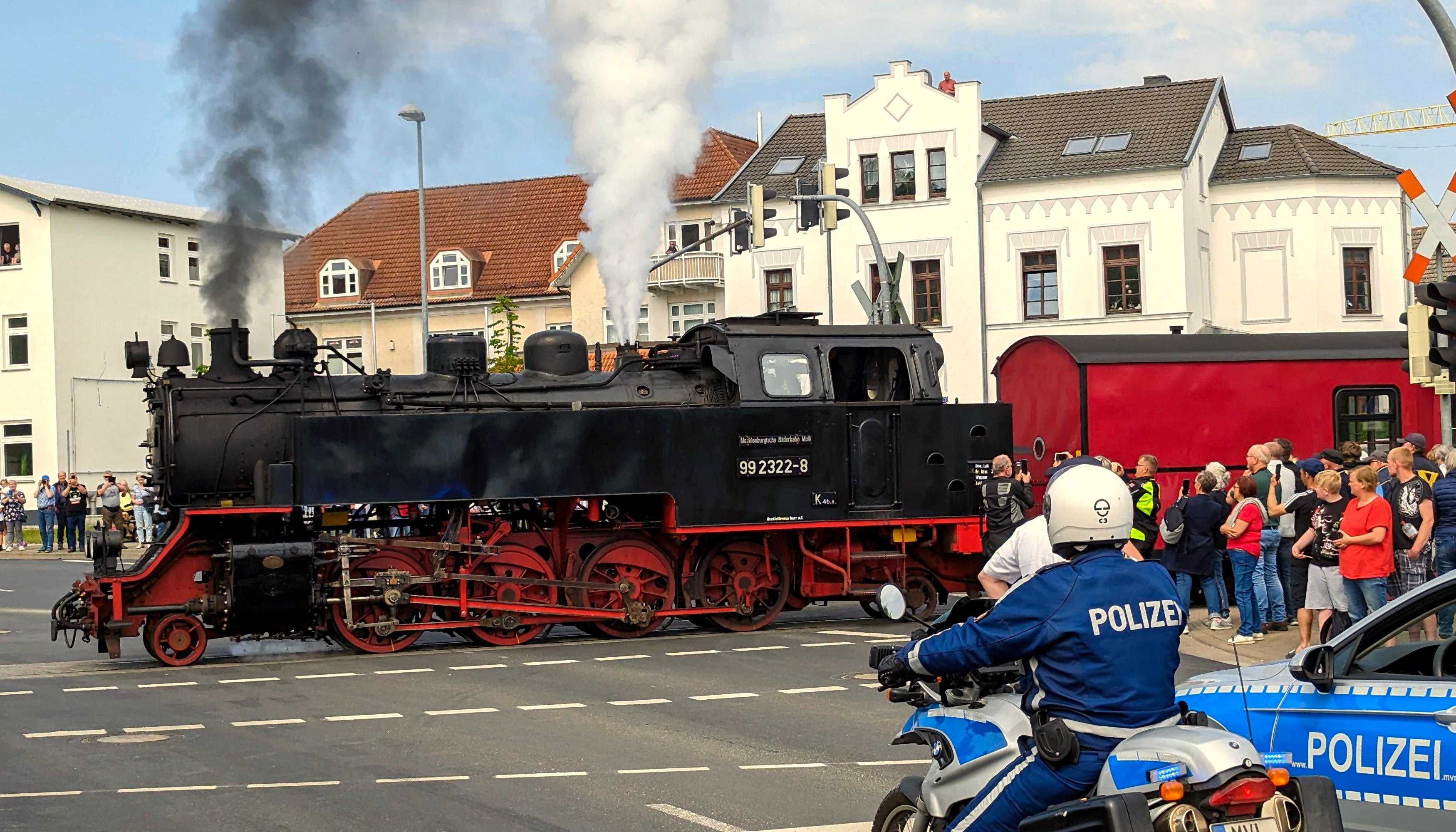 Das Bild zeigt eine Dampflokomotive, die gerade einen Zug über eine Kreuzung zieht. Aus ihrem Schornstein steigt Rauch auf, aus den weiter hinten angeordneten Pfeifen Dampf. Die Lokomotive trägt die Nummer 99 3222-8. 
An den Gleisen stehen viele Menschen. Auf der Straße ein Polizeiauto und ein Polizist auf einem Motorrad.
Hinter dem Zug sind Häuser zu sehen. 
Blauer Himmel und weiße Wölkchen.