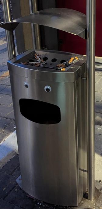 Ein Mülleimer mit Kulleraugen über dem Einwurf/Mund und Aschenbecher oben.