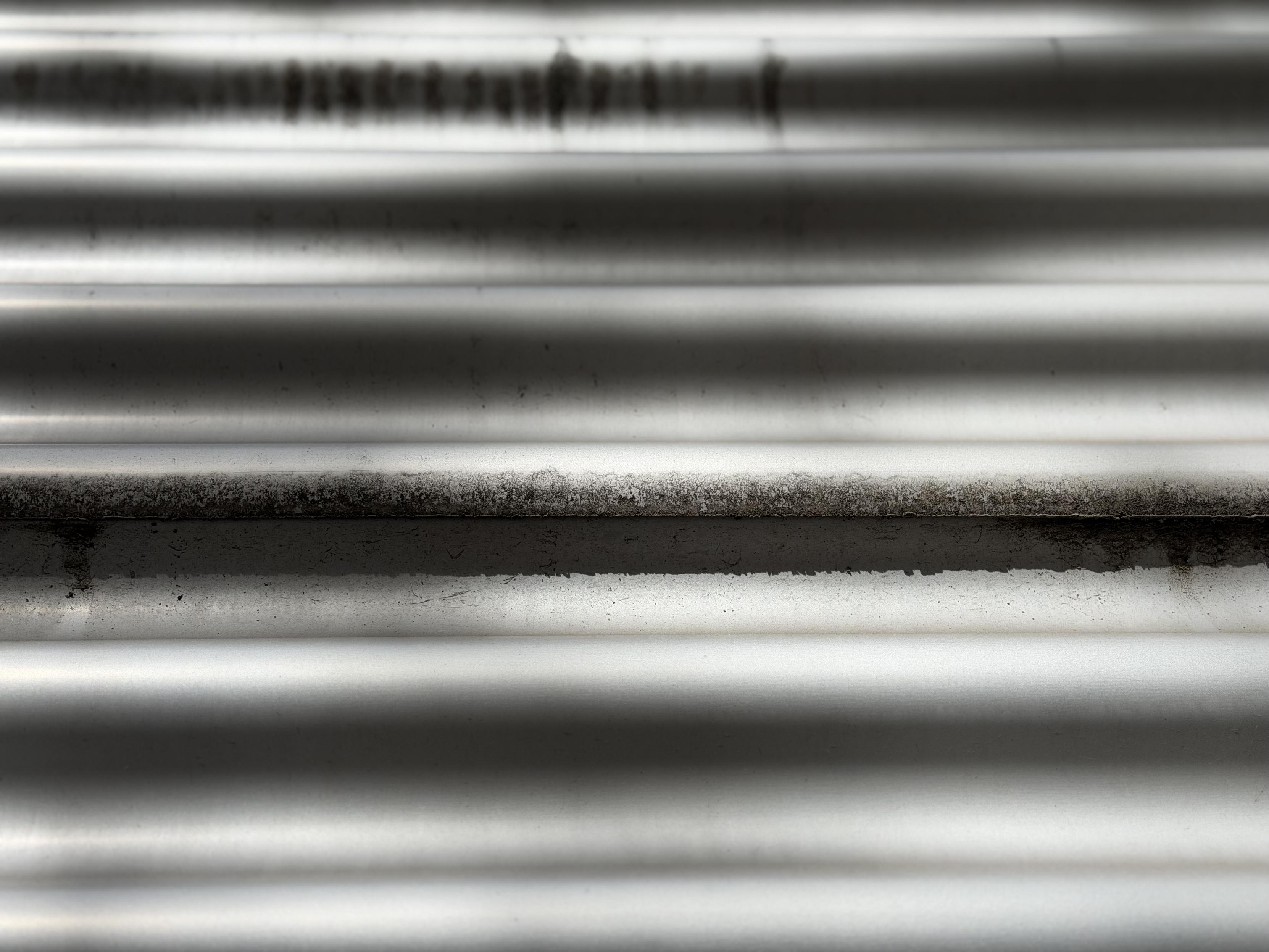 Nahaufnahme einer horizontal gewellten Metalloberfläche, vermutlich Wellblech, in hellem Grau bis Silber. Die Rillen verlaufen parallel über das gesamte Bild und werfen durch das Streiflicht abwechselnd helle und dunkle Streifen. Entlang einer der oberen Rillen ist eine unscharfe, dunkle Musterung zu erkennen, die wie eine Beschriftung oder ein Aufdruck wirkt. An mehreren Stellen, besonders im mittleren Bildbereich, haften dunkle, krümelige Verschmutzungen oder Ablagerungen an den erhabenen Kanten.