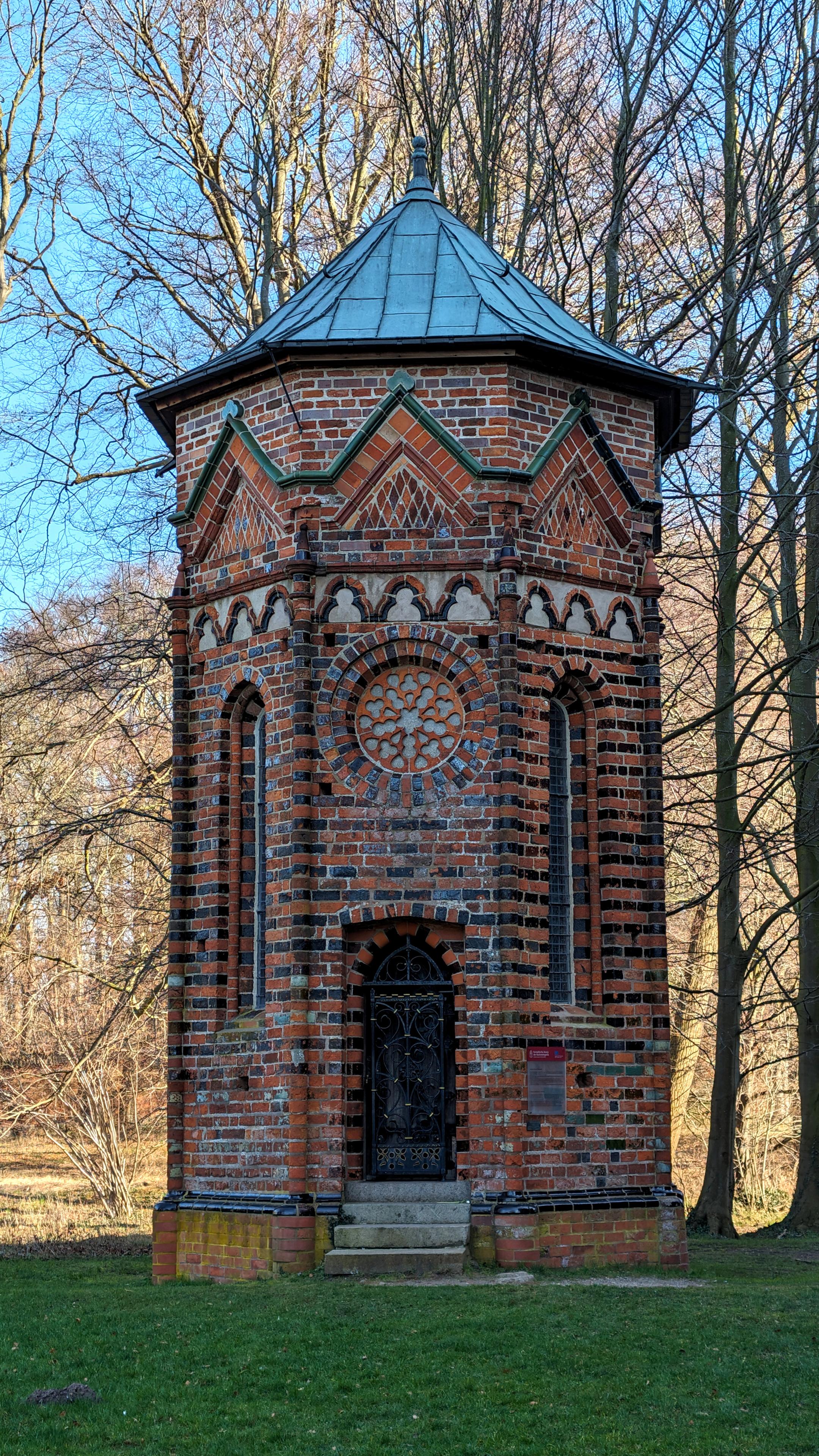 Das Bild zeigt einen turmartigen, achteckigen Bau aus roten und glasierten schwarzen Backsteinen, der auf einer Rasenfläche steht. 
Die Fassade, die zum Betrachter zeigt, enthält eine schwarze Holztür, zu der vier Stufen hinaufführen. Über der Tür ein rundes Ornament aus Backstein. Darüber ein dreiteiliges Kleeblattfries. Die anderen Seiten des Gebäudes sehen ähnlich aus, haben aber anstelle der Tür und dem Ornament schmale Fenster mit Rundbögen.
Ein Kupferdach schließt den Turm nach oben ab. 
Im Hintergrund kahle Bäume und blauer Himmel.