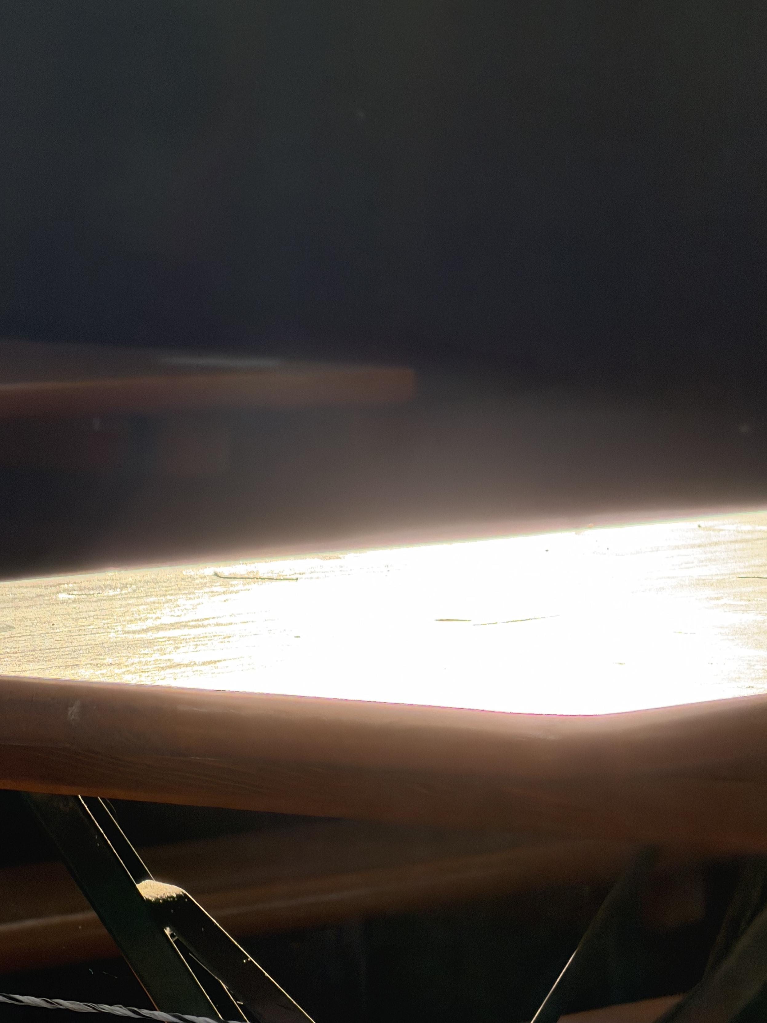 Kontrastreiche Nahaufnahme einer Holzkante einers Biergartentisches, auf die grelles Sonnenlicht fällt und dabei eine überstrahlte, fast weiße Fläche erzeugt. Der obere Bildbereich liegt vollständig im Dunkeln, nur die untere Kante der Holzstruktur ist deutlich sichtbar mit warmem, bräunlichem Ton. Im unteren linken Bildbereich sind dünne, sich kreuzende Metallstreben zu erkennen.