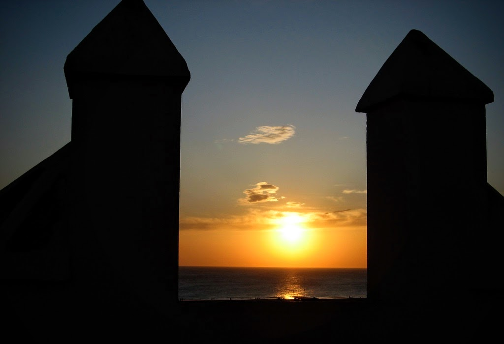 Die dunklen Silhouetten der Zinnen einer Wehrmauer rahmen einen goldgelben Sonnenuntergang über dem Meer. Die Sonne steht knapp über dem Horizont und spiegelt sich auf dem Wasser. Am Himmel sind wenige kleine Wolken zu sehen.