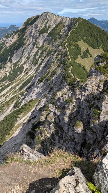 Blick von einem Gipfel auf den nächsten in der Kette, auf den ein schmaler Bergpfad führt und der sich hell vom Grün und der Felsen abzeichnet. Steile Abgründe an der Nordseite links des Pfades und Geröllfelder. Sonne steht im Mittag.
