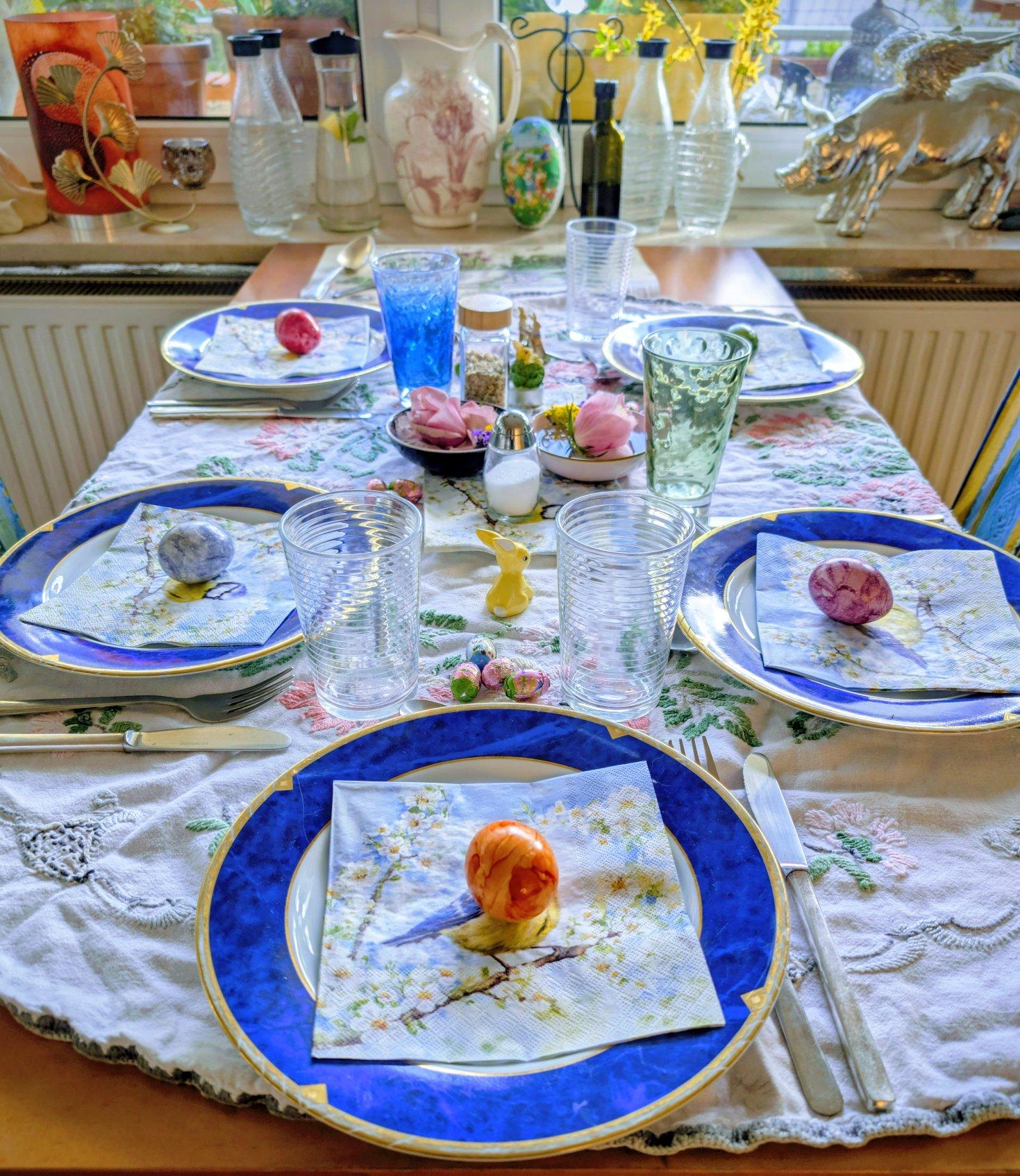 Ein gedeckter Tisch in einem Wohnzimmer. Auf dem  Tisch liegt eine weiße Tischdecke mit Blumenmuster. Auf 5 Tellern liegen Servietten mit Blaumeisen drauf, auf den Servietten Ostereier. Zwischen den Tellern Osterhasen aus Porzellan. Im Hintergrund auf einem Fensterbrett Flaschen, Vasen und ein geflügeltes Schwein.