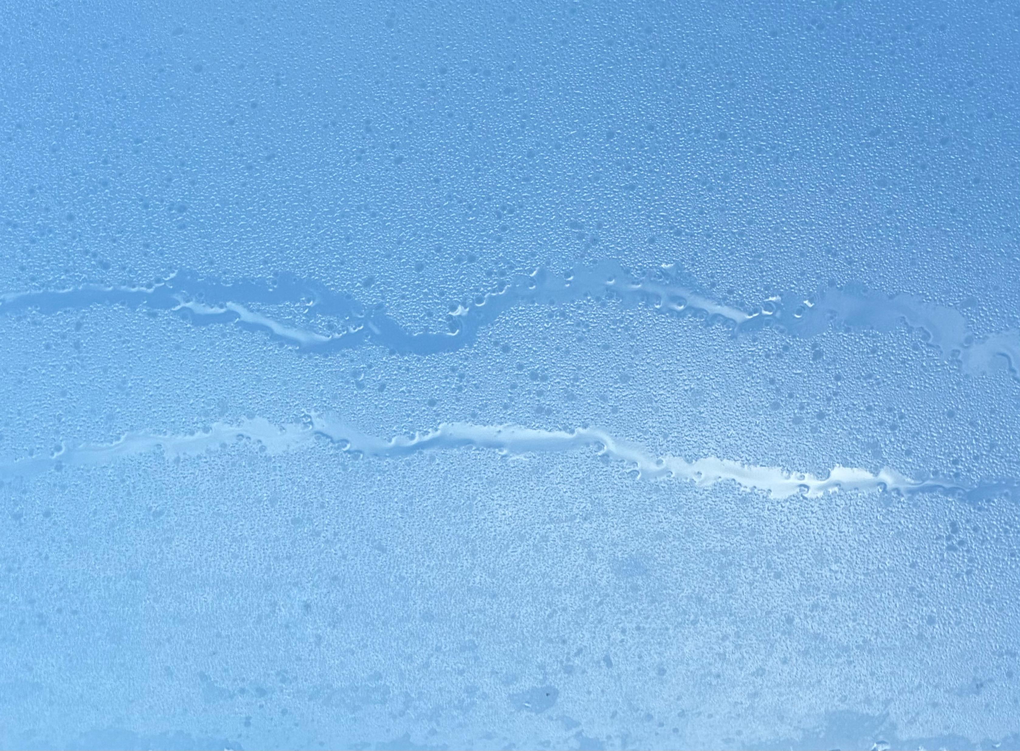 Nahaufnahme einer mit feinen Kondenstropfen bedeckten Glasfläche gegen den Himmel. Zwei horizontale, geschwungene Linien mit größeren, zusammengelaufenen Wassertropfen ziehen sich parallel durch das Bild und heben sich als klare, glänzende Bahnen von der übrigen, gleichmäßig feuchten Oberfläche ab.