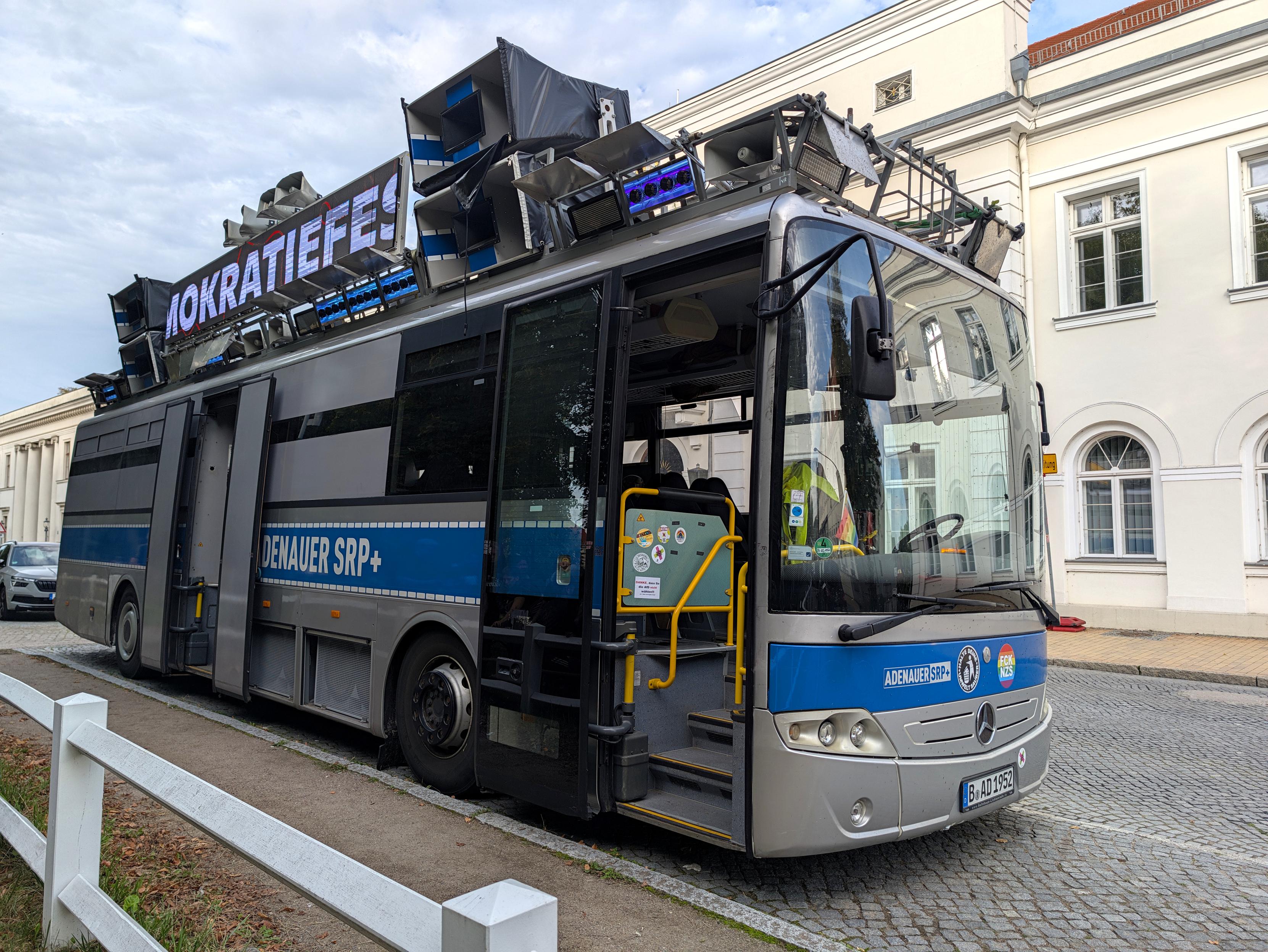 Auf dem Seitenstreifen einer gepflasterten Straße steht ein silberfarben lackierter Bus mit blauen Aufklebern im Polizeistil. Darauf steht "ADENAUER SRP+". Auf dem Dach des Fahrzeugs sind große Lautsprecher eine Anzeige für Laufschriften und Strahler für Lichteffekte installiert.
Im Vordergrund ein weiße Holzzaun.
Auf der anderen Straßenseite hinter dem Bus ein repräsentatives klassizistisches Gebäude mit weißer Fassade.