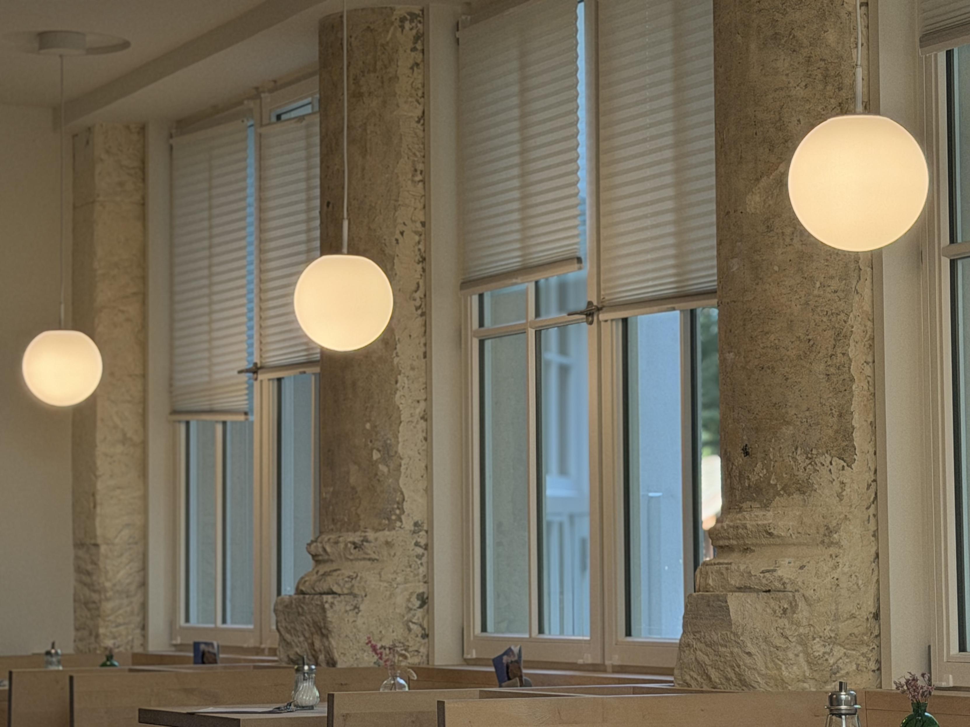 Innenaufnahme des Cafés im Alpines Museum in München mit hohen Sprossenfenstern, teils mit heruntergelassenen Plisseerollos. Drei kugelförmige, weiß leuchtende Pendelleuchten hängen von der Decke, unterschiedlich hoch und in Reihe angeordnet. Zwischen den Fenstern stehen massive, grob verputzte Sandsteinpfeiler mit profilierten Basen. Im unteren Bildbereich sind hölzerne Tischgruppen mit Salz- und Pfefferstreuern sowie kleinen Blumenvasen zu erkennen.
