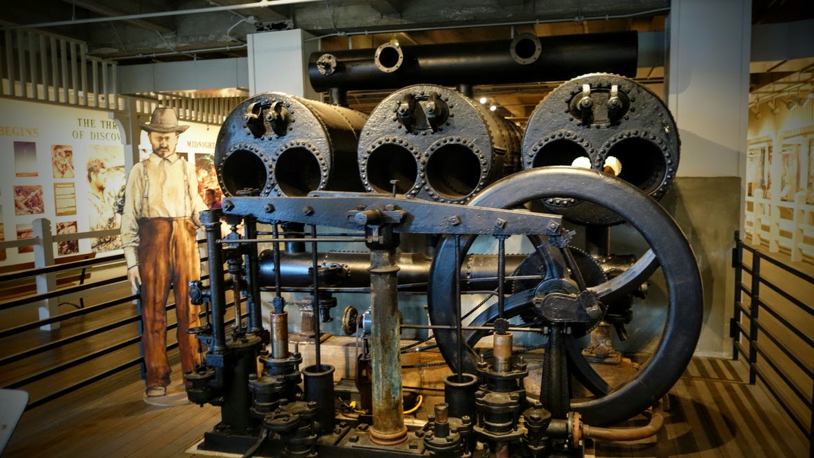 Historische Dampfmaschine in einem Museum. Eine große schwarze, genietete Maschine mit mehreren runden Zylindern, Rohren, Ventilen, Stangen und einem großen Schwungrad steht hinter einer Absperrung. Links daneben ist eine lebensgroße Darstellung eines Arbeiters zu sehen.