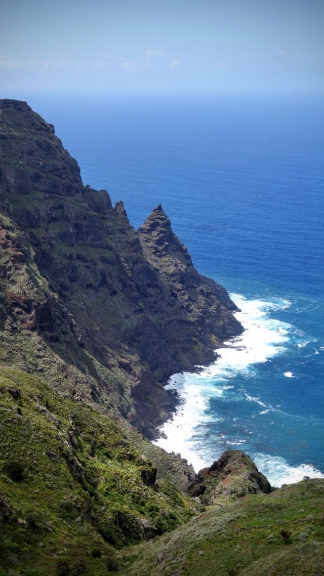 Steile, grün bewachsene Felsküste am tiefblauen Meer. Links ragen dunkle, zerklüftete Klippen ins Bild, dazwischen ein spitzer Felsvorsprung. Unten brechen weiße Wellen an den Felsen. Im Hintergrund erstreckt sich das ruhige, weite Meer bis zum leicht dunstigen Horizont unter einem hellblauen Himmel.