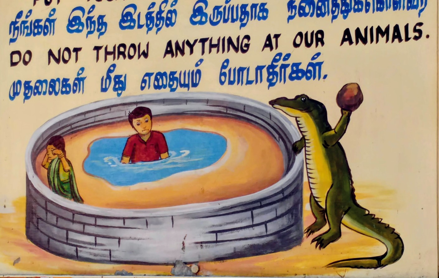 Ein Schild mit einer Zeichnung und Text in tamilischer und englischer Sprache und Schrift: Do not throw anything at our animals. (Werfe nichts auf unsere Tiere). 
Darunter ist eine Zeichnung zu sehen, auf der zwei Kinder (ein Junge und ein Mädchen) sich in einem runden Gehege, umgeben von einer Mauer befinden. Der Junge sitzt in in der Mitte des Geheges in einem Pool mit Wasser, das Mädchen links am Rand, es weint. Rechts am Rand steht ein Krokodil außerhalb der Mauer und wirft einen großen Stein auf die Kinder innerhalb der Mauern.