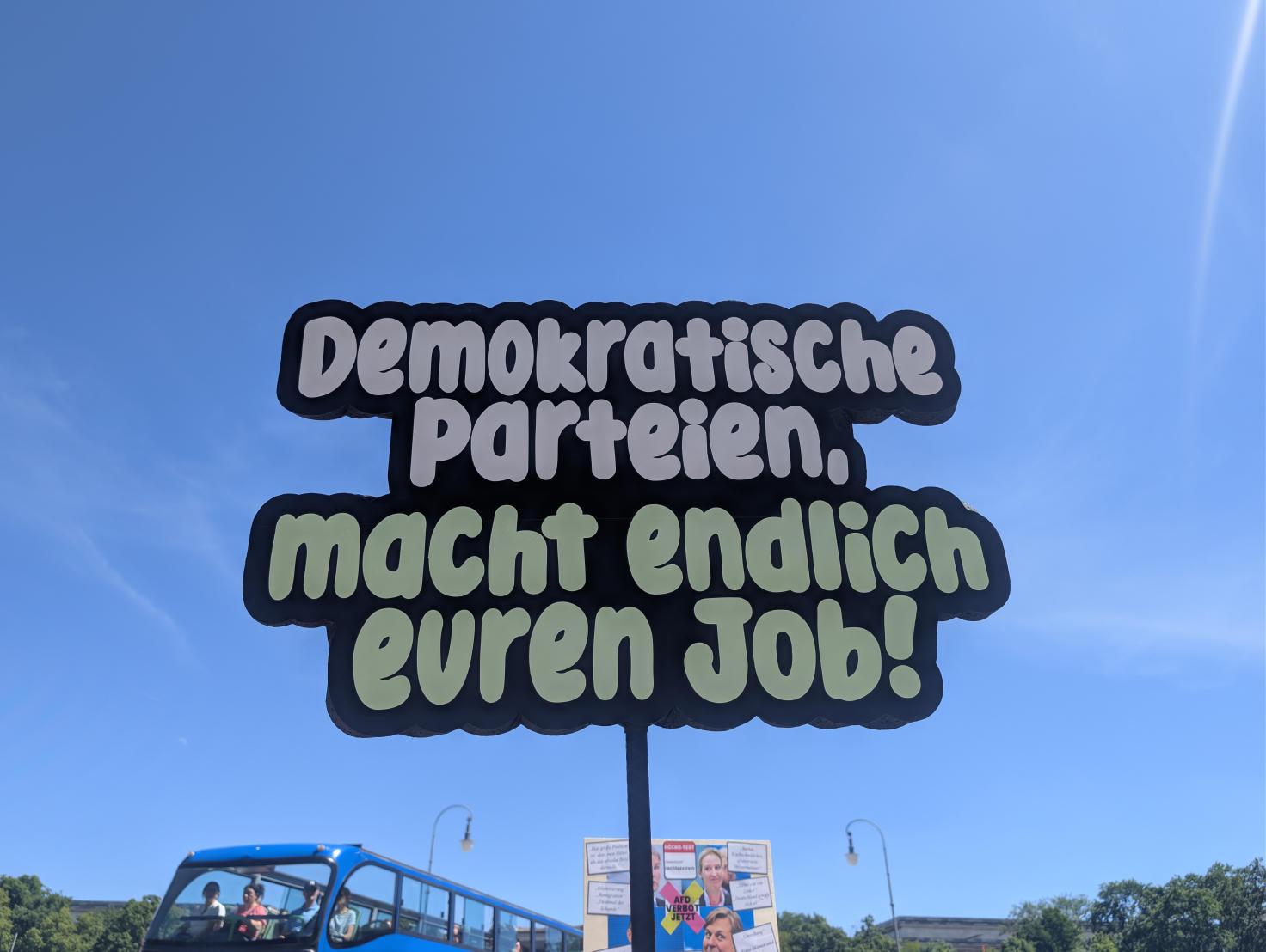 Ein Schild aus Holz auf einer Demo, zu einer Sprechblase zugeschnitten: Demokratische Parteien, macht endlich euren Job!
Dahinter ein blauer Himmel, darunter links ein Touristenbus mit offenem Verdeck, rechts ein weiteres Demoschild.