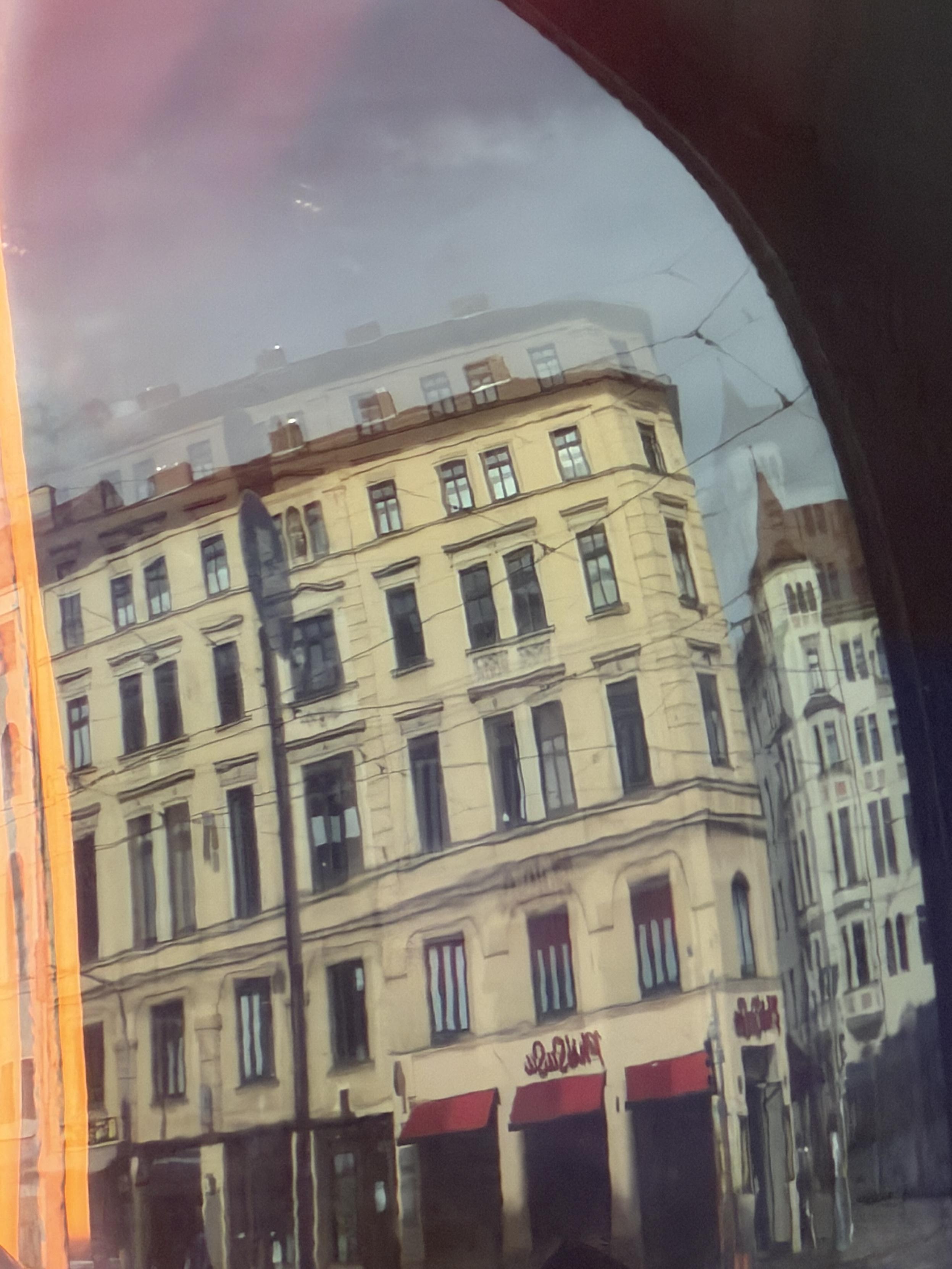 Durch eine gebogene, leicht rissige Glasscheibe fotografierte Straßenszene mit einem hellgelben, historischen Eckgebäude mit mehreren Fensterreihen und roten Markisen im Erdgeschoss. Am unteren Rand des Gebäudes ist Graffiti-Schrift zu erkennen. Rechts im Hintergrund ein weiteres, weißes Gebäude mit Türmchen. Der obere Bildrand zeigt bewölkten Himmel, an den seitlichen Rändern verläuft ein orangefarbener Lichtreflex, rechts oben verdunkelt eine gebogene, dunkle Fläche den Bildrand.