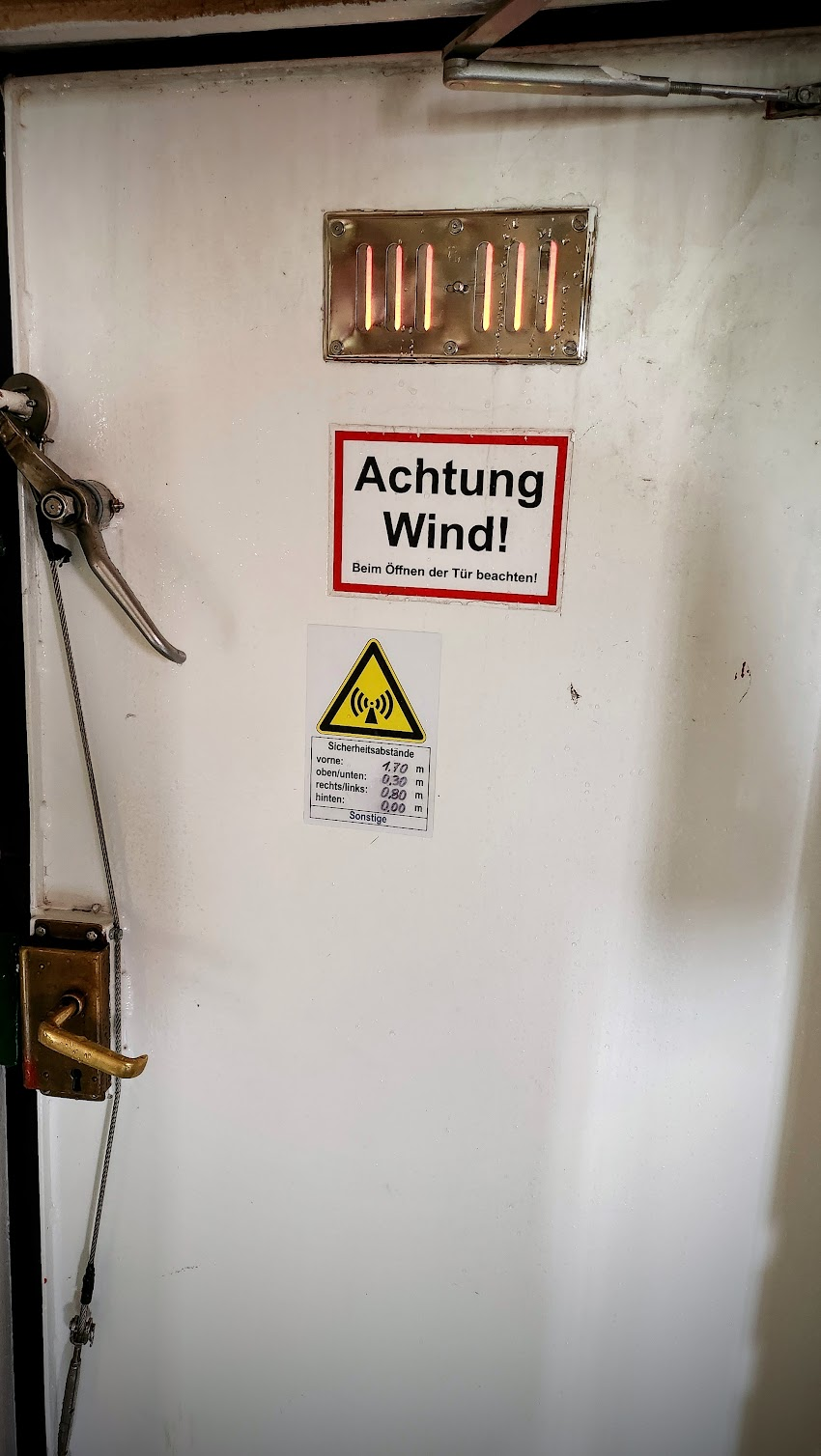 Weiße, metallene Tür in einem technischen Innenraum. Links befinden sich ein mechanischer Türverschluss und ein Messing-Türgriff mit Seilzug. Oben an der Tür ist ein kleines Lüftungsgitter. Darunter hängen zwei Warnschilder: Das obere trägt die Aufschrift „Achtung Wind! Beim Öffnen der Tür beachten!“. Das darunter zeigt ein gelbes Warnsymbol für Funkwellen und nennt Sicherheitsabstände. Die Tür wirkt robust und industriell.