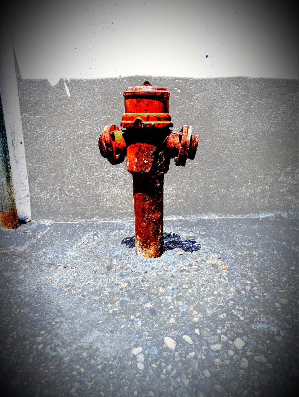 Ein roter Hydrant aus Metall, etwas altertümlich anmutend, auf dem Gehweg, dahinter eine Hausmauer.