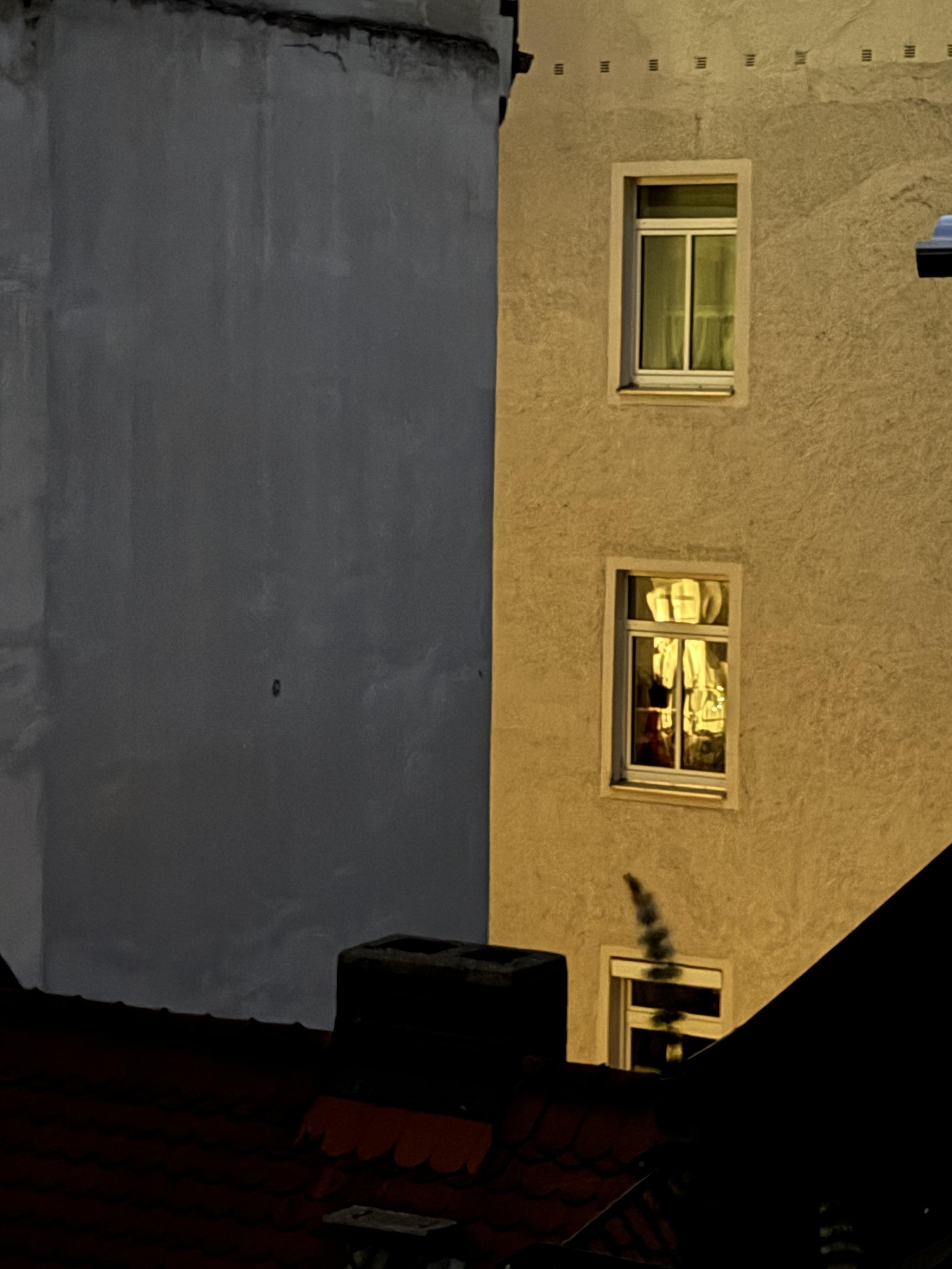Nächtliche Aufnahme zweier aneinandergrenzender Hausfassaden. Links eine glatte, graue Wand, rechts eine golden-warm beleuchtete Fassade mit zwei weiß gerahmten Sprossenfenstern übereinander – im oberen Fenster hängt ein heller Vorhang, im unteren sind Gegenstände und Stoffbahnen erkennbar. Darunter ein drittes, dunkleres Fenster, vor dem sich eine unscharfe, dunkle Form abzeichnet. Am unteren Bildrand liegt im Schatten ein rotes Ziegeldach mit einem quaderförmigen Schornstein.