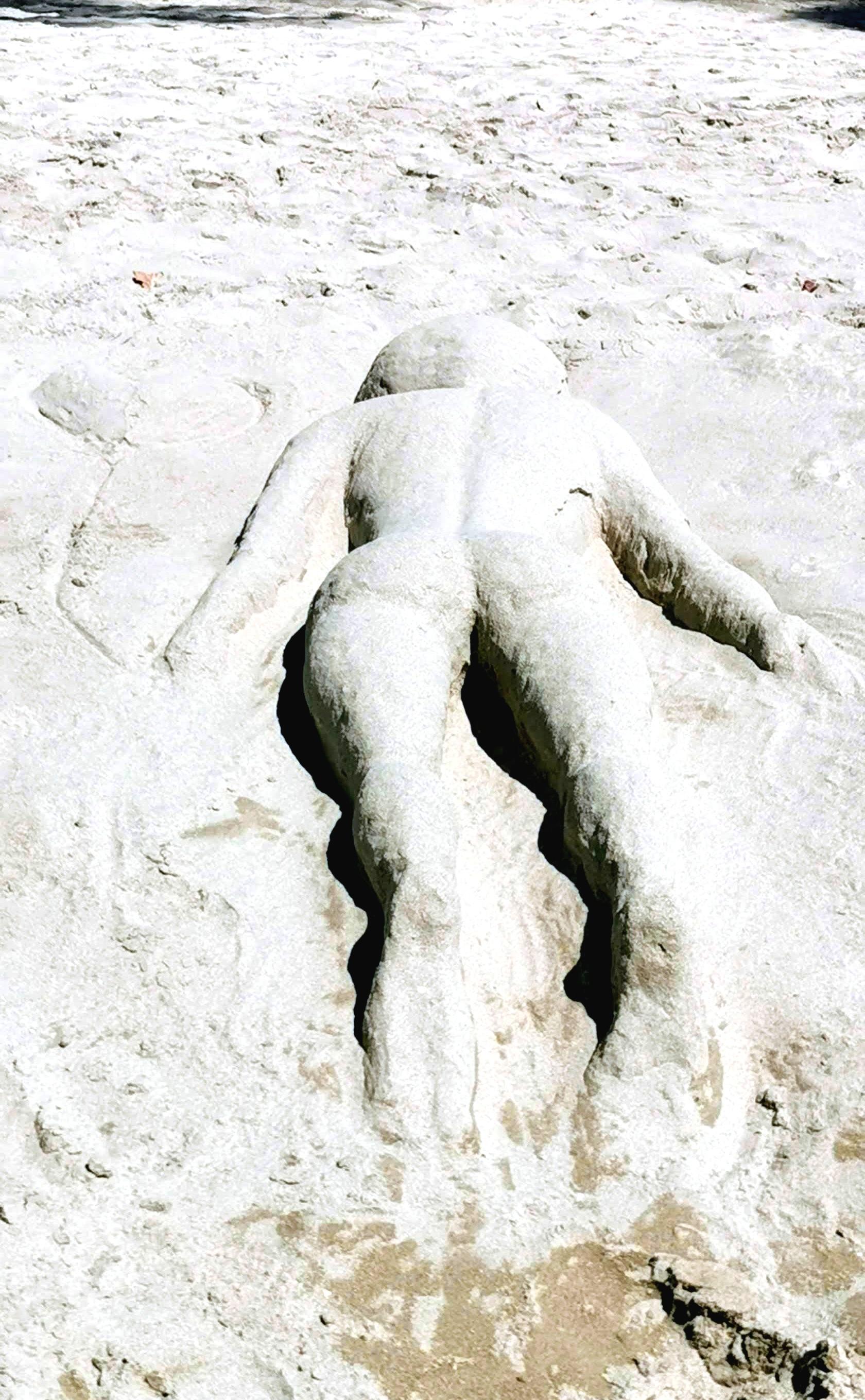 Foto an einem Strand. Aus dem Sand ist eine liegende Frauenfigur gestaltet worden.Sie liegt auf dem Bauch im Sand, das Rückgrat, der Po, Beine und Füße sind gut zu erkennen.