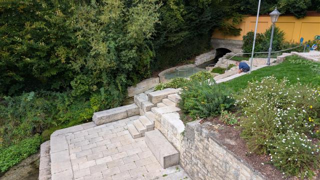 Aus einer Öffnung unter einer gelben Mauer fließt Wasser in ein Kneippbecken und danach weiter in die Altmühl unterhalb davon. Davor sind Steinstufen und Sitzgelegenheiten  aus hellgrauen Steinquadern angelegt. Rechts daneben ein Blumenbeet, dahinter Höhe Bäume.